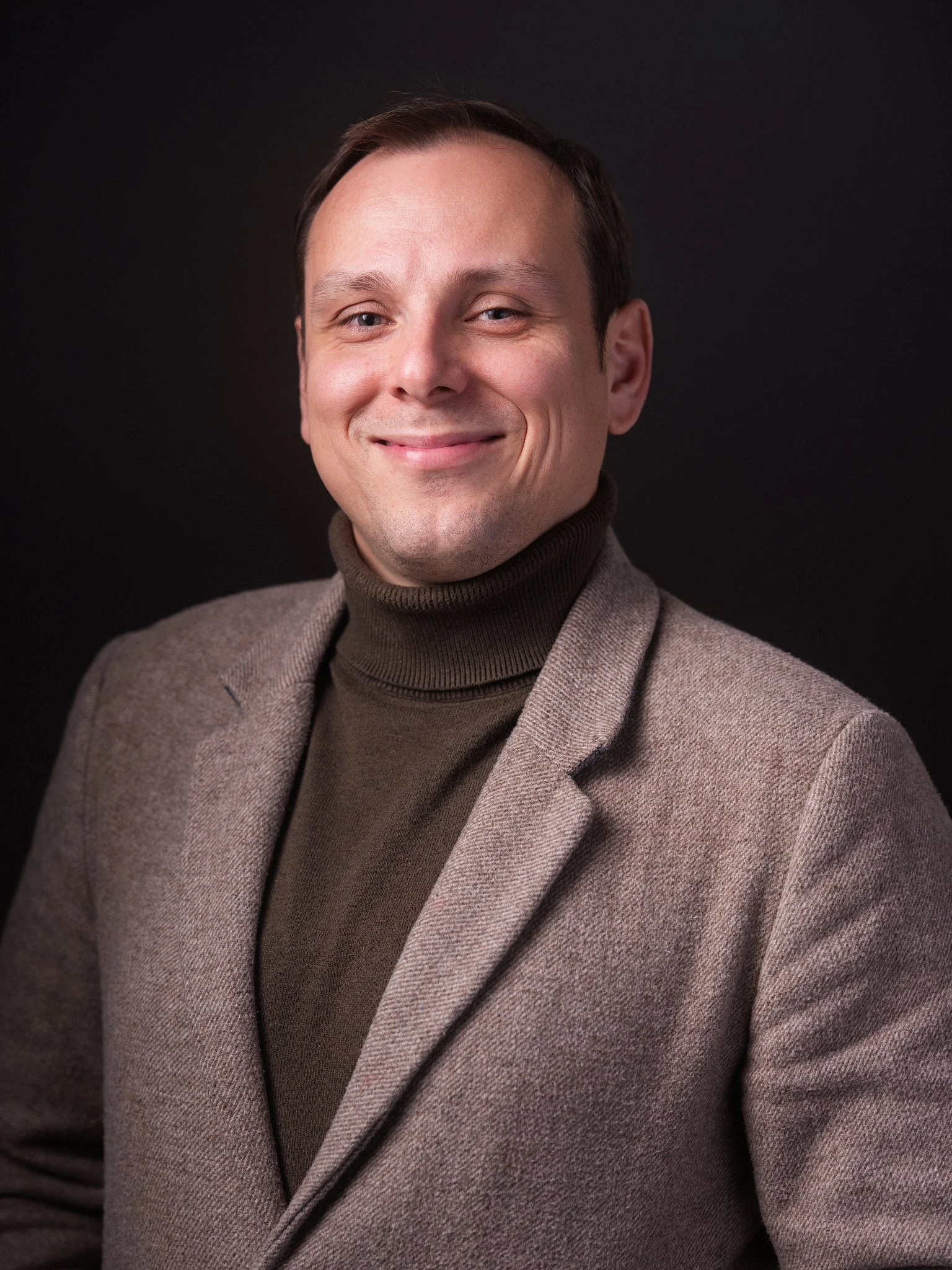 A man with brown hair and a beige sweater looking intently at a laptop with his finger on his chin in a coffee shop or cafe.