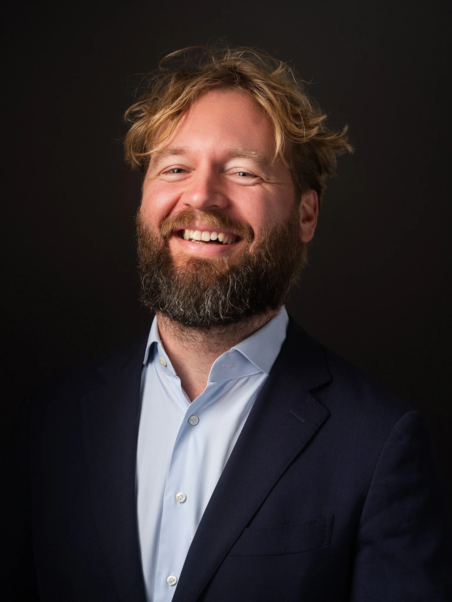 A man with a beard and mustache smiling, wearing a black button-up shirt, with a background of green plants and a window with a wooden frame.