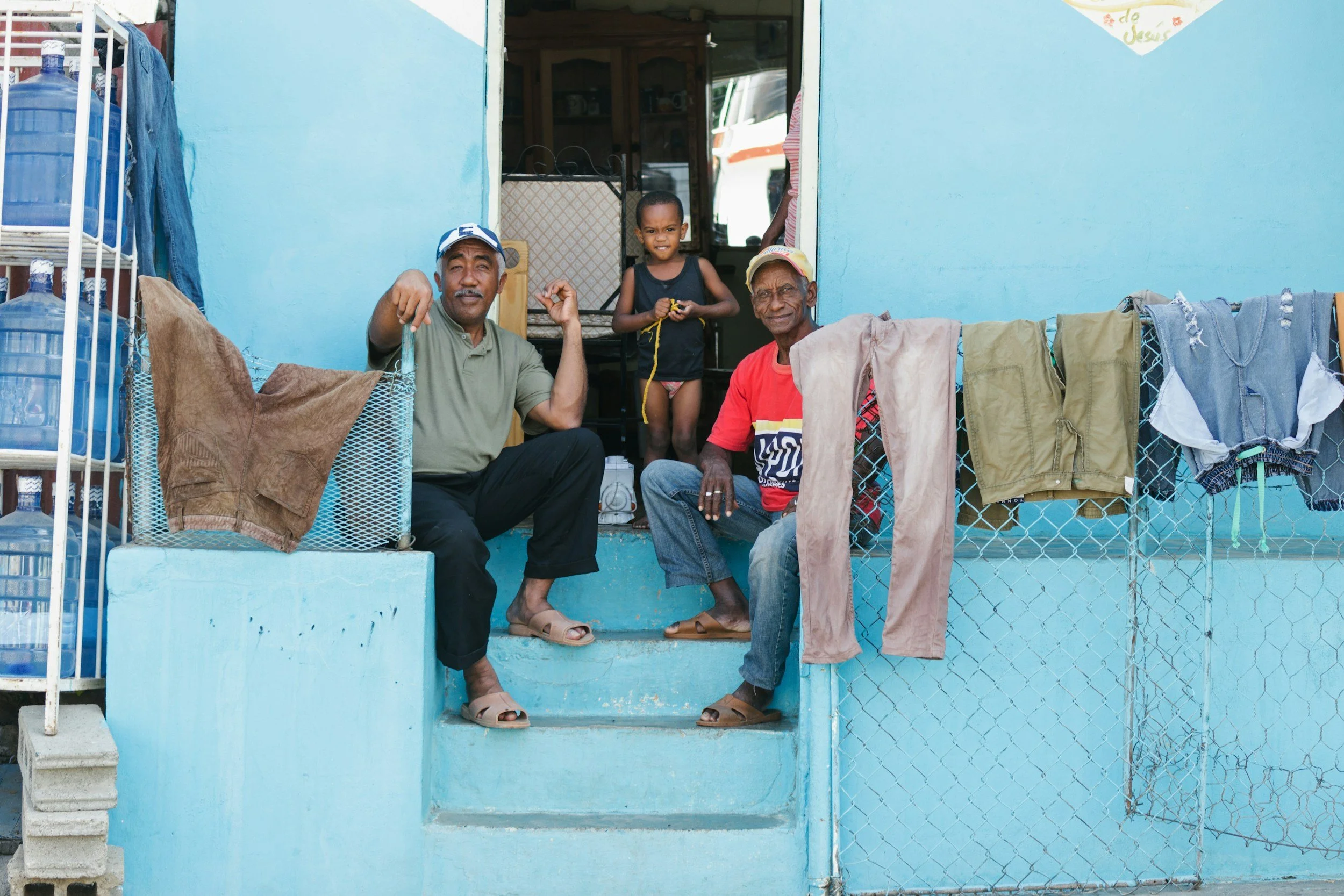 Two elderly men and a young boy sitting on the steps of a blue house, with laundry hanging on the fence, and the men smiling at the camera.