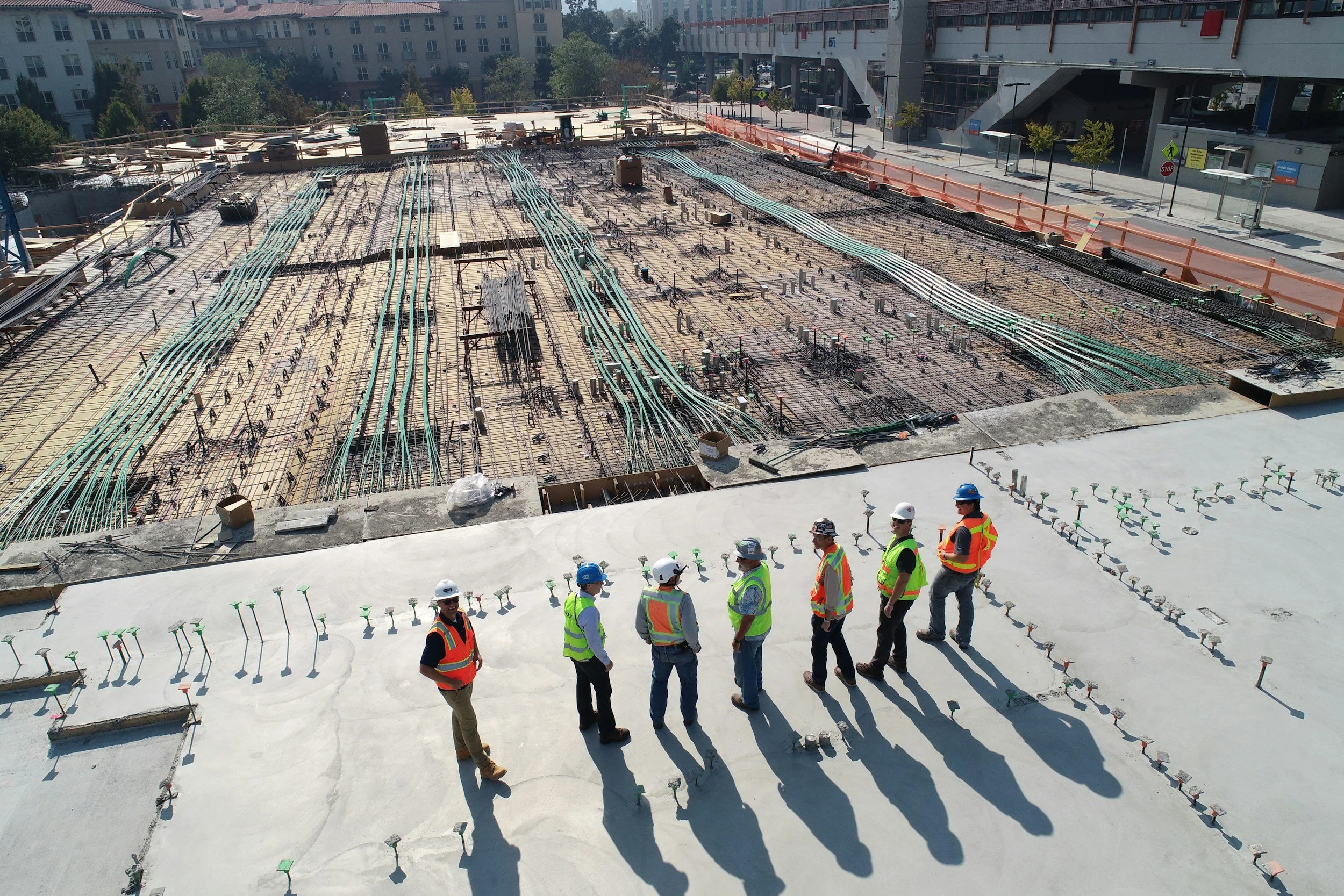 Construction workers in safety gear standing on a concrete surface at a building site, with an unfinished construction area featuring exposed rebar and numerous electrical conduits and piping in the background.