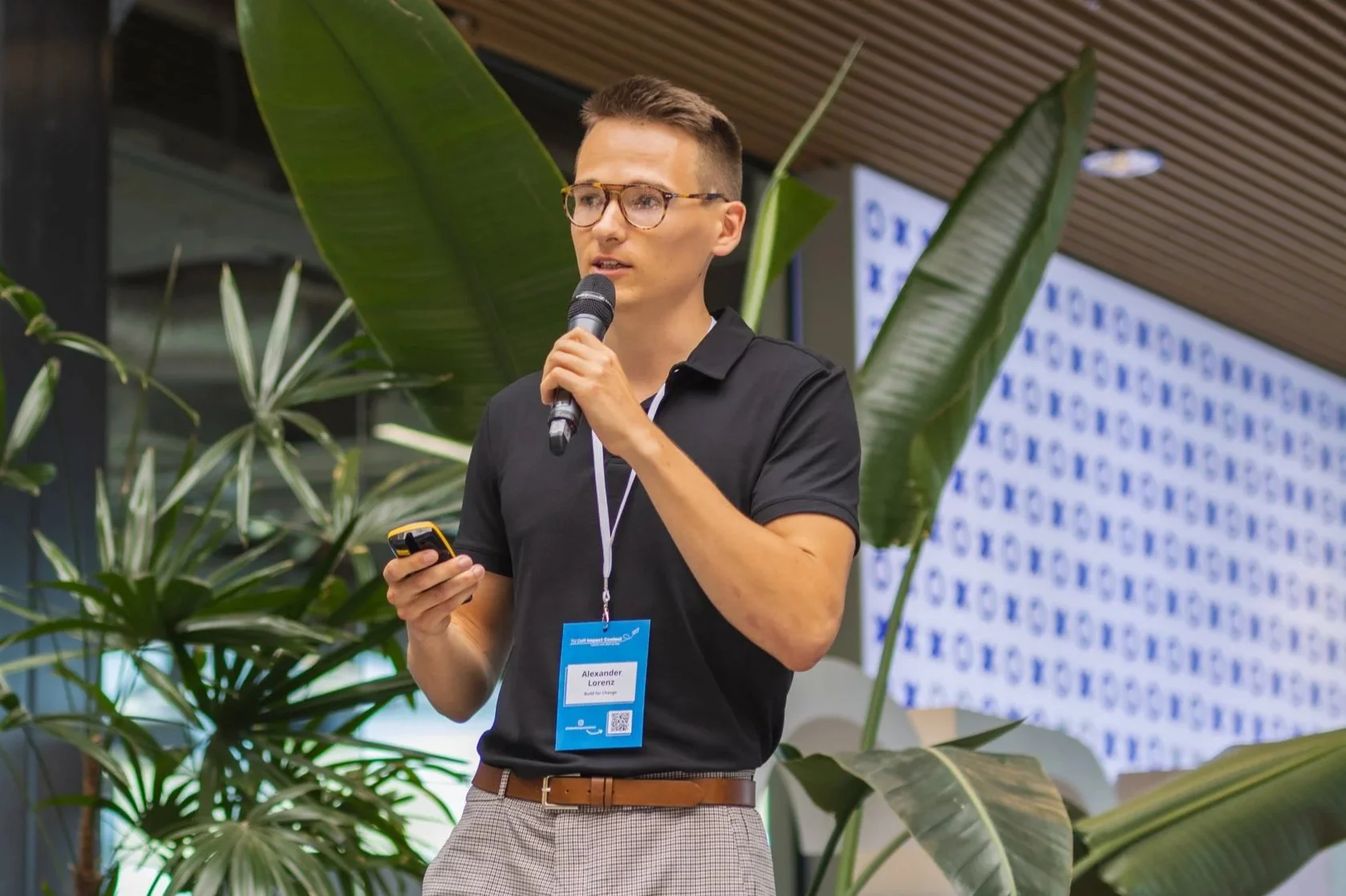A man holding a microphone and a smartphone, standing among large green plants and speaking at an event with a blue and white patterned background.