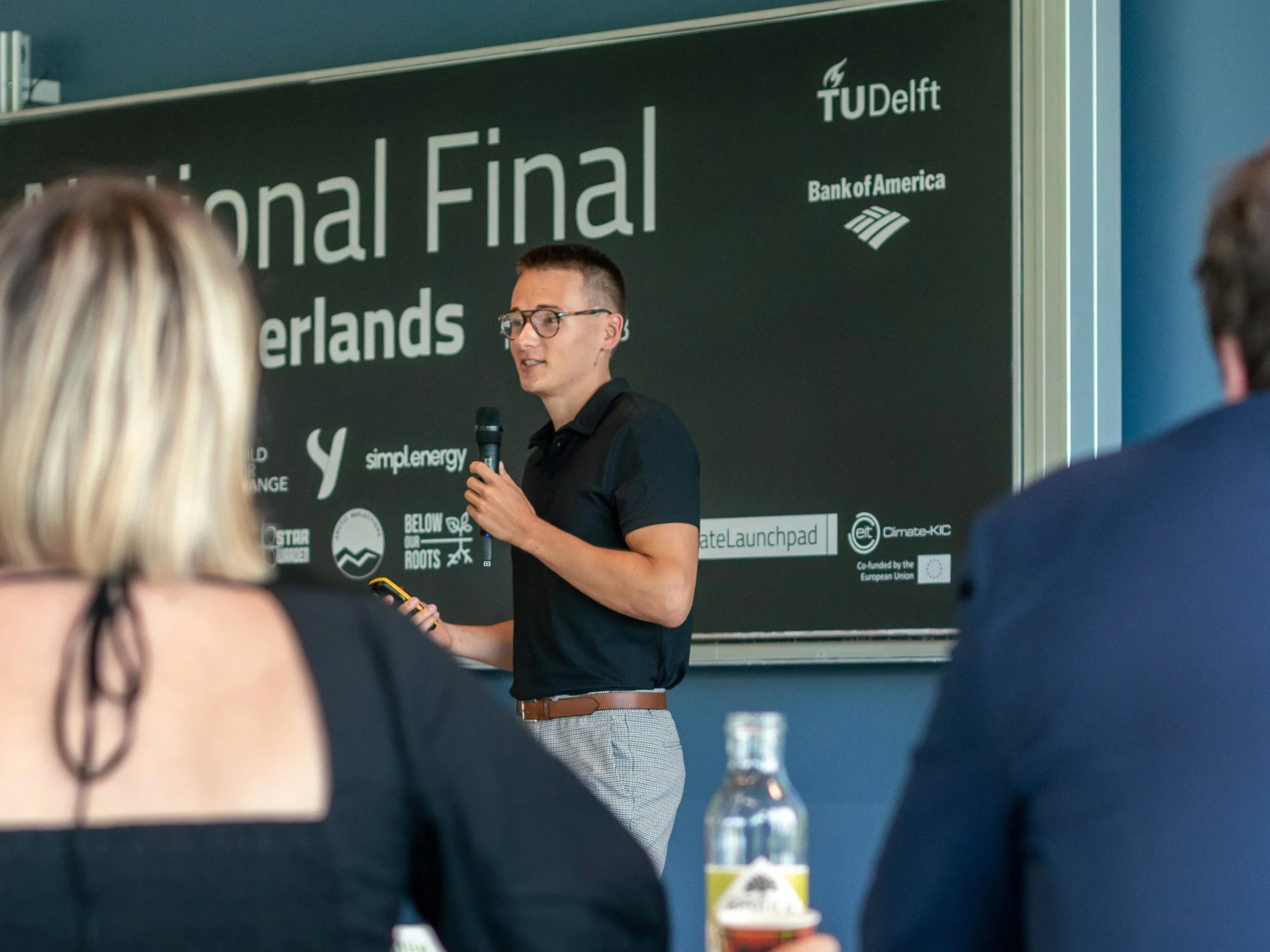 A young man with glasses presenting at a conference, holding a microphone and a remote control, with a dark green backdrop that reads 'National Final' and contains various sponsor logos.