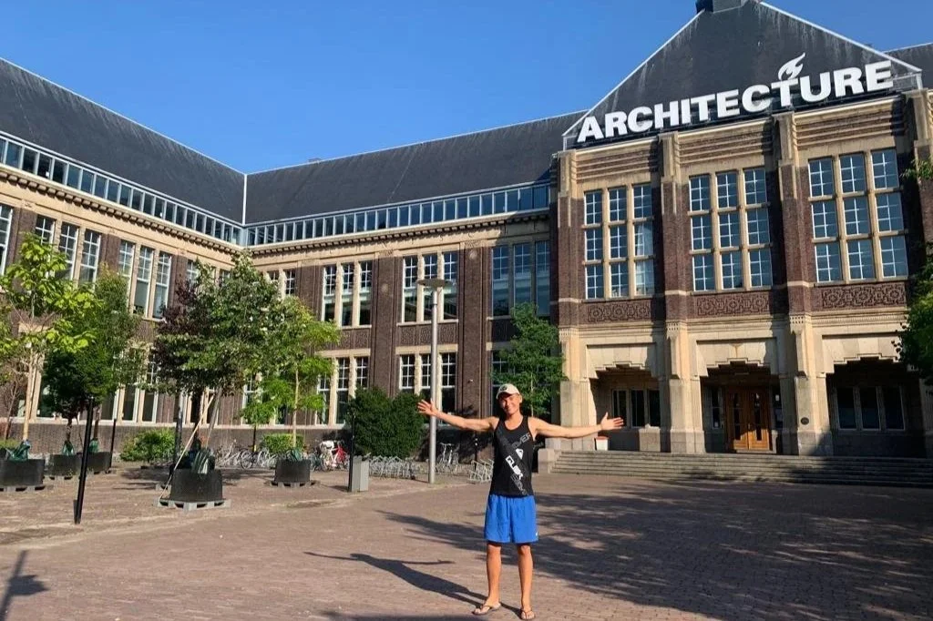 A person in a black tank top, blue shorts, and a baseball cap stands with arms outstretched in front of a large brick building with a sign that reads "ARCHITECTURE." The building has multiple large windows and decorative architectural details. There are trees, bikes, and outdoor seating in the plaza under a clear blue sky.