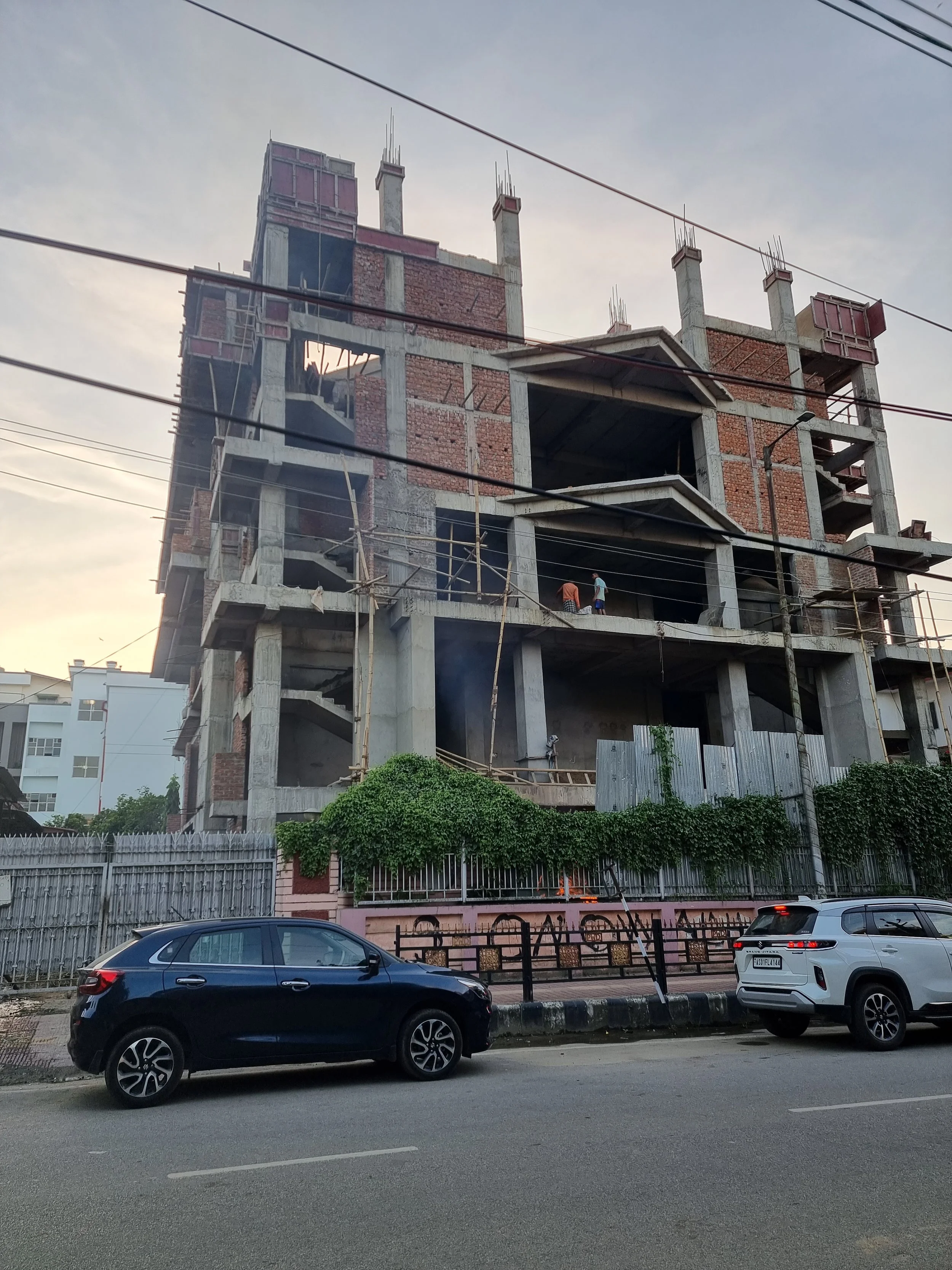 A multi-story building under construction with visible concrete framework and brick walls, located on a city street with parked cars in the foreground.