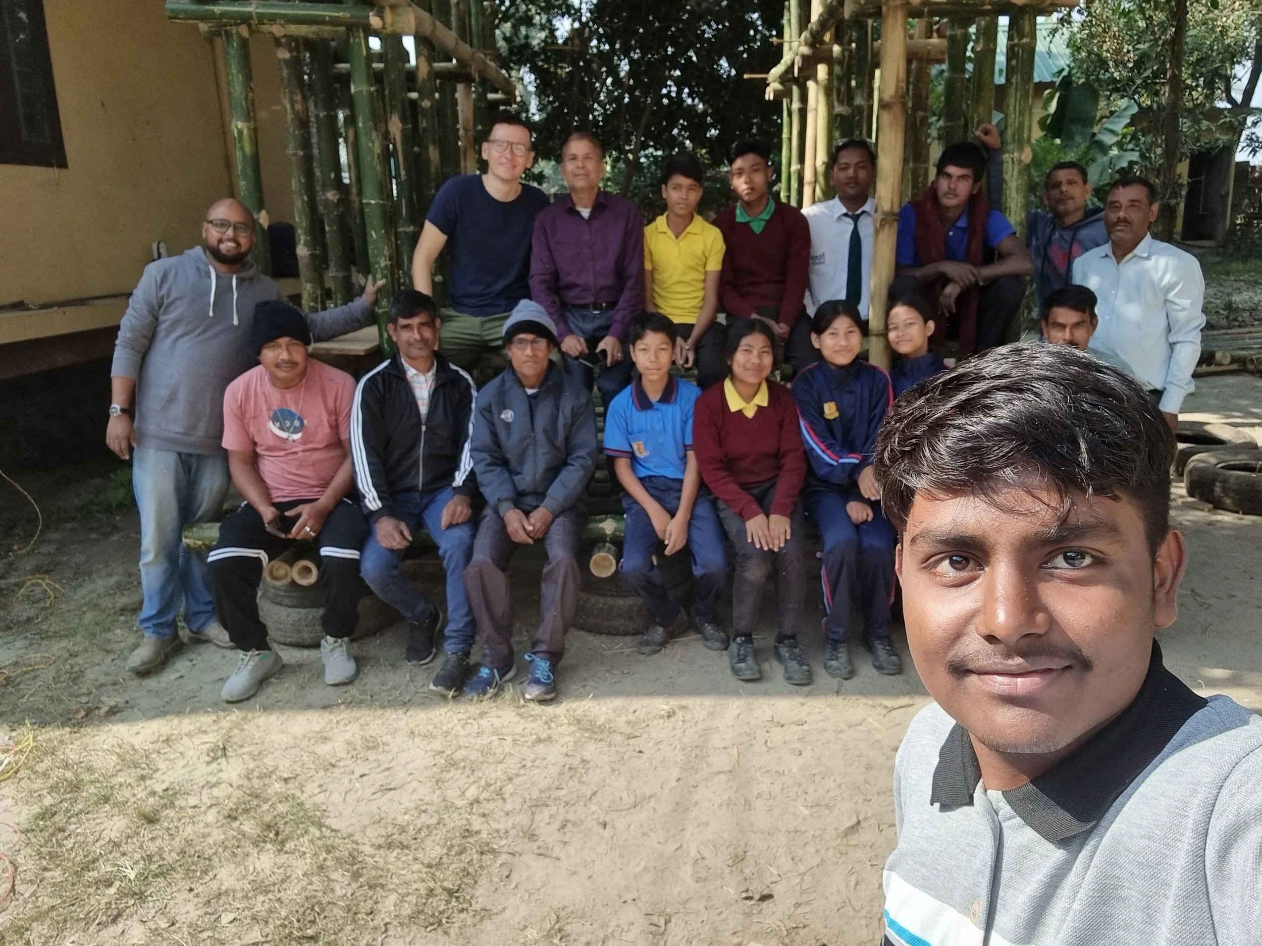 A group of people, including children and adults, posing outdoors for a photo. Some are sitting on a tire bench, while others are standing behind them. The background has bamboo and greenery, and the ground is dirt.