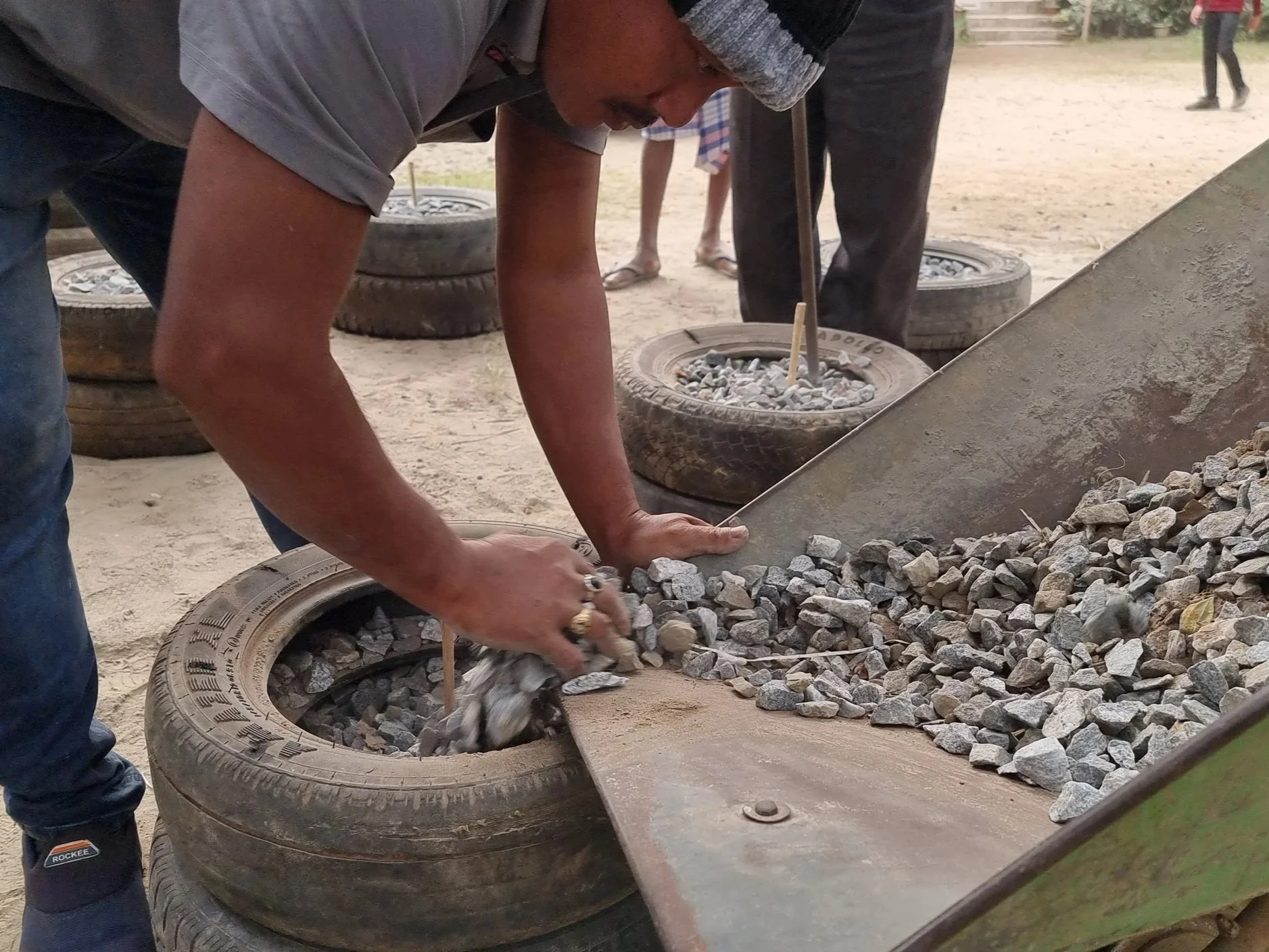 A man working with rocks on a makeshift conveyor made of car tires and metal in an outdoor setting with a dirt ground.