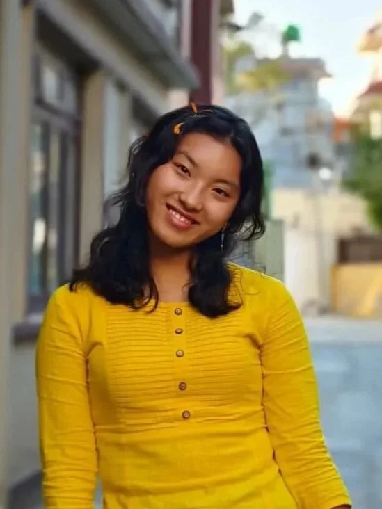 Smiling young woman with black wavy hair, wearing a yellow long-sleeve shirt, standing outdoors in a residential area.