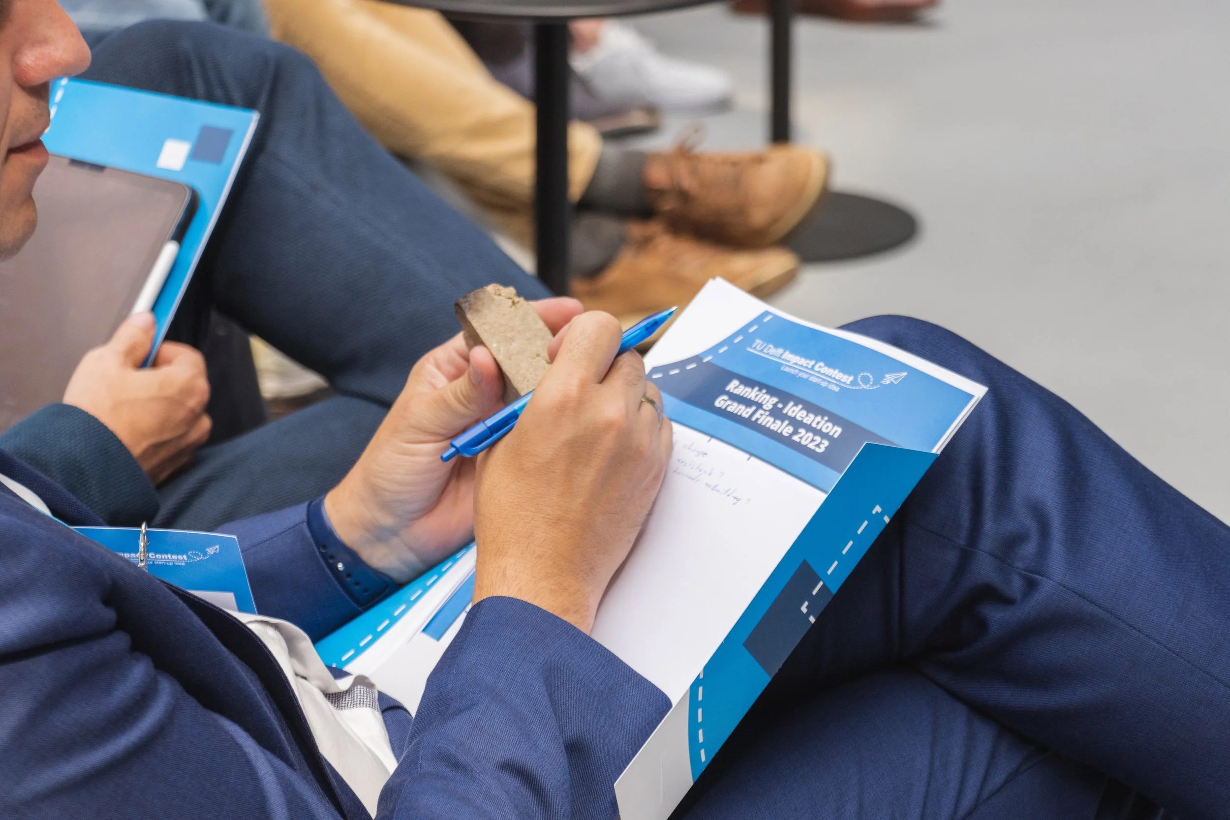 Person in a blue suit writing on a notepad at a conference, with a brochure titled 'Ranking - Ideation Grand Finale 2023' on their lap, and another person holding a phone nearby.