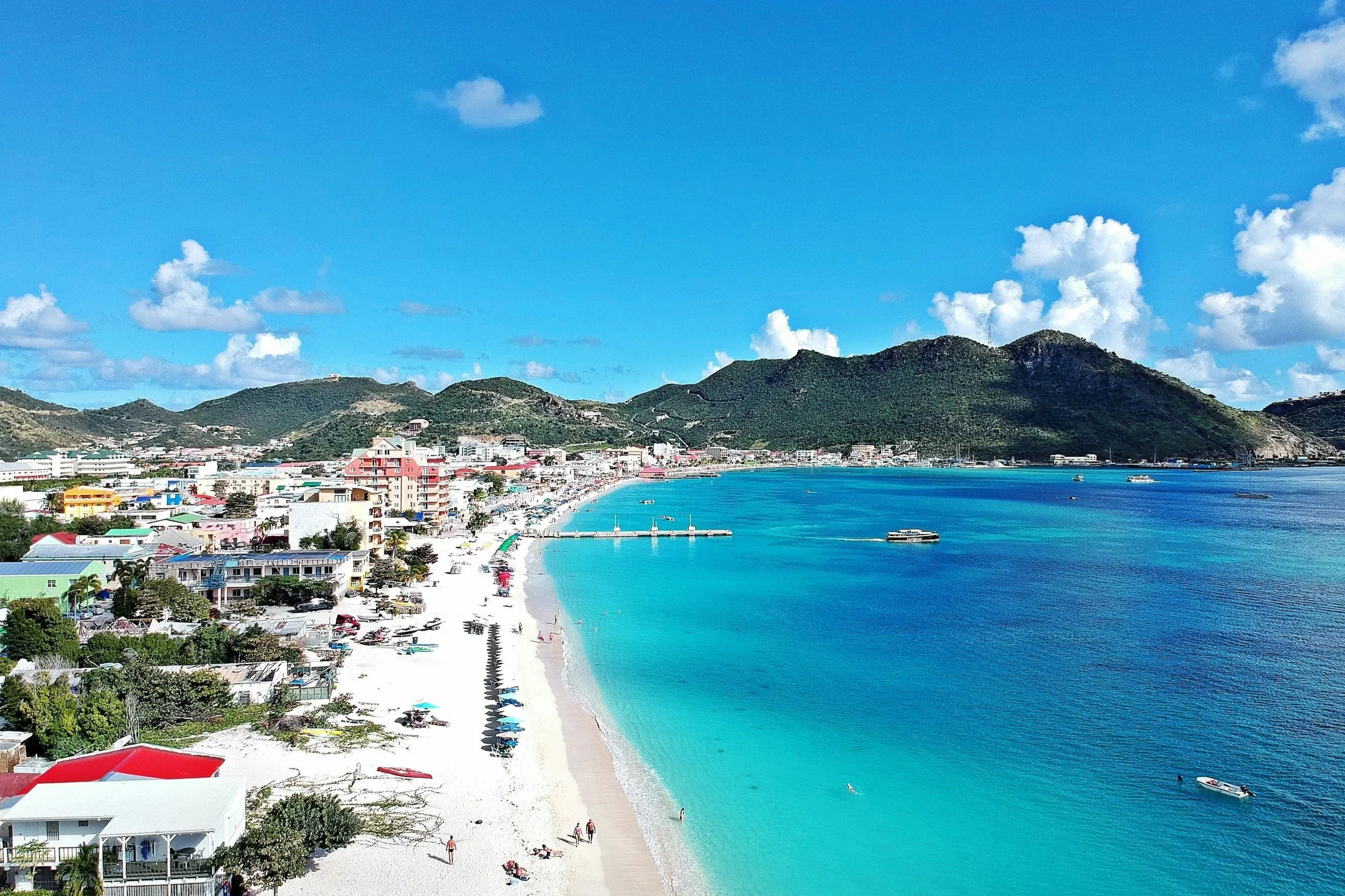 A vibrant beach scene with turquoise water, a white sandy shoreline, beach umbrellas, and a town with colorful buildings against green hills under a partly cloudy sky.