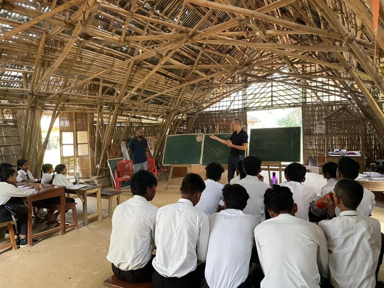 A classroom inside a building made of bamboo, with students sitting on benches and listening to a teacher at the blackboard, with another person standing near the teacher.