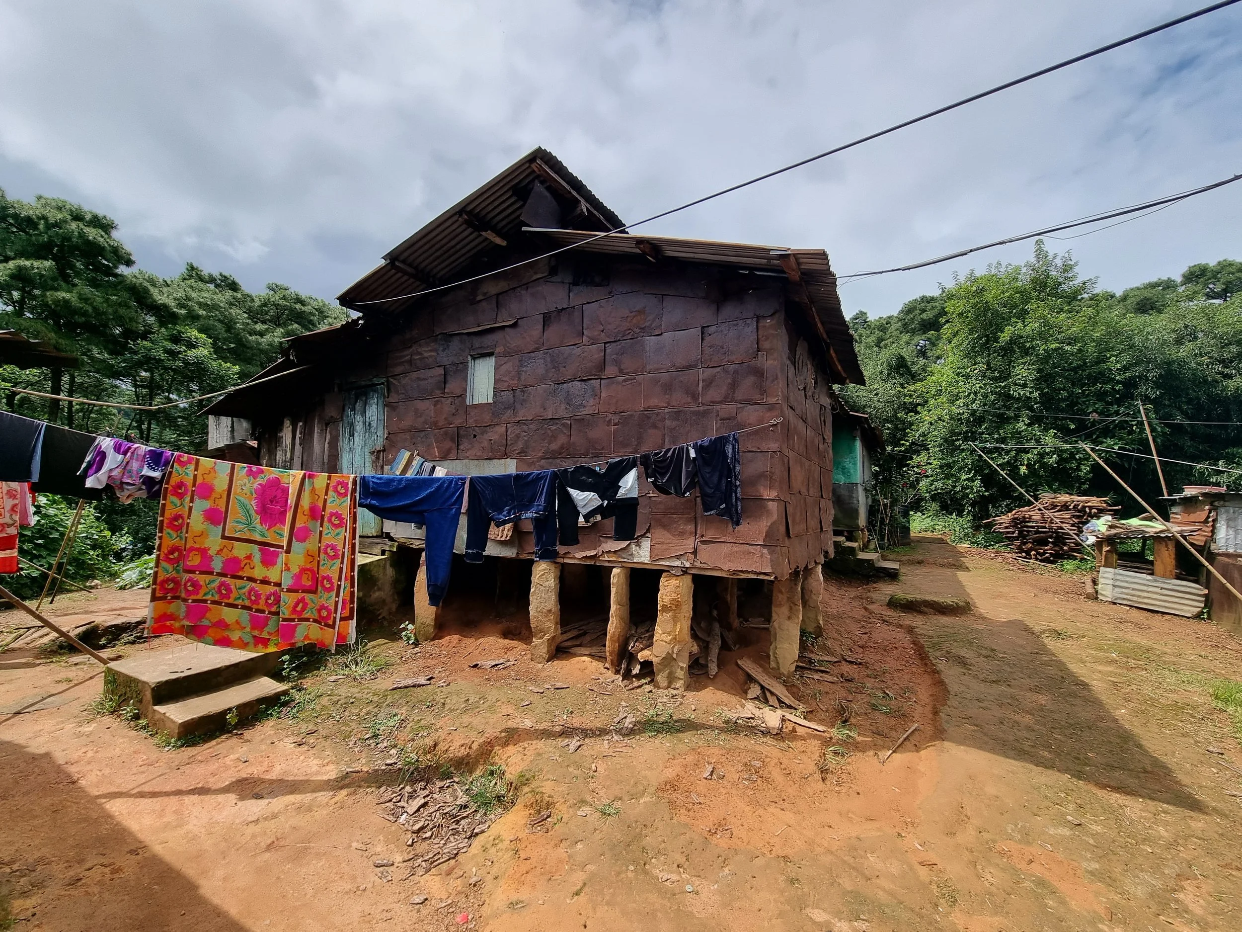 A rustic house elevated on stone pillars in a rural area with trees and laundry hanging outside.