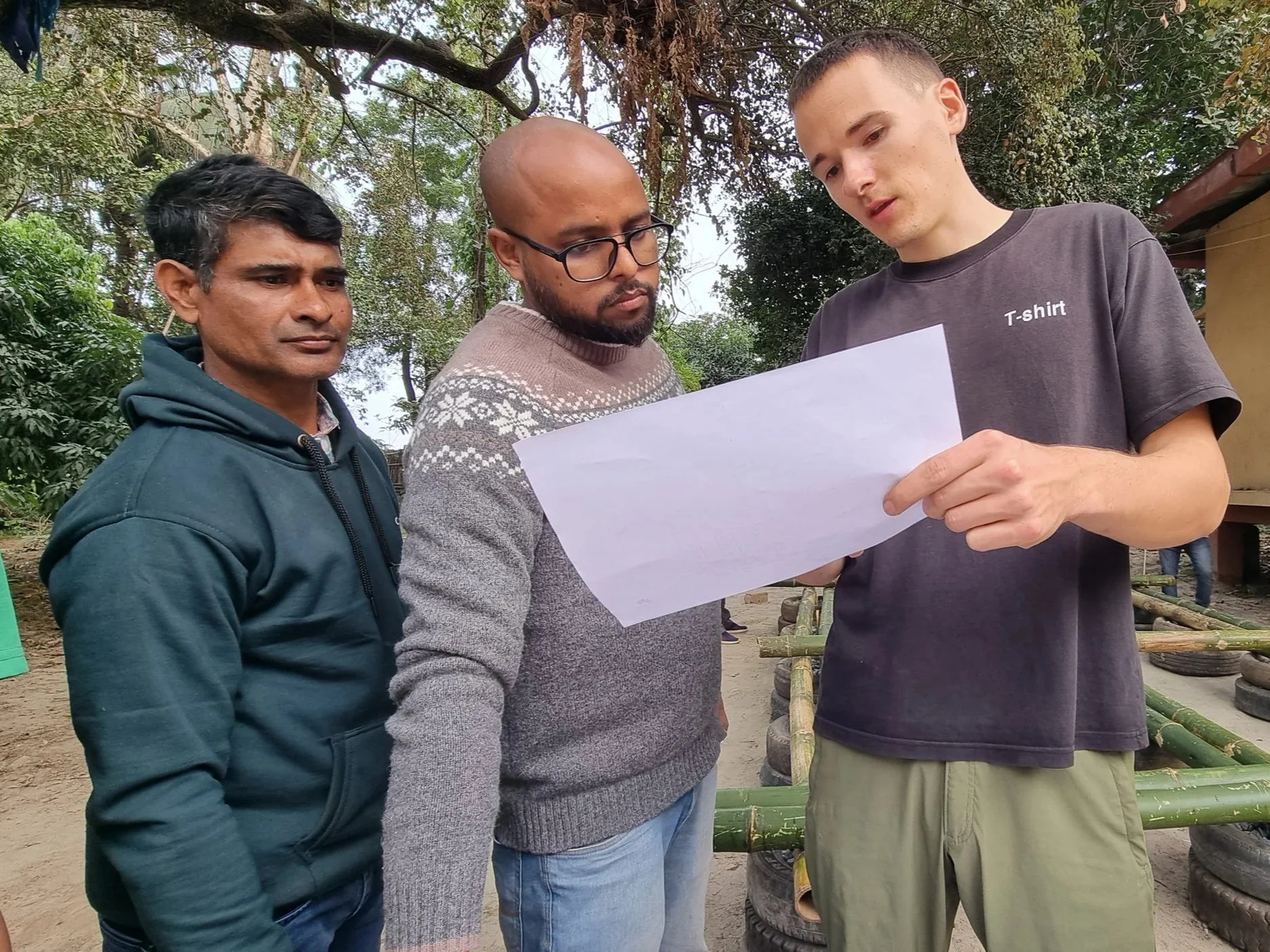 Three men standing outdoors, looking at a piece of paper held by the man on the right. The background includes trees and a structure made of tires and bamboo.
