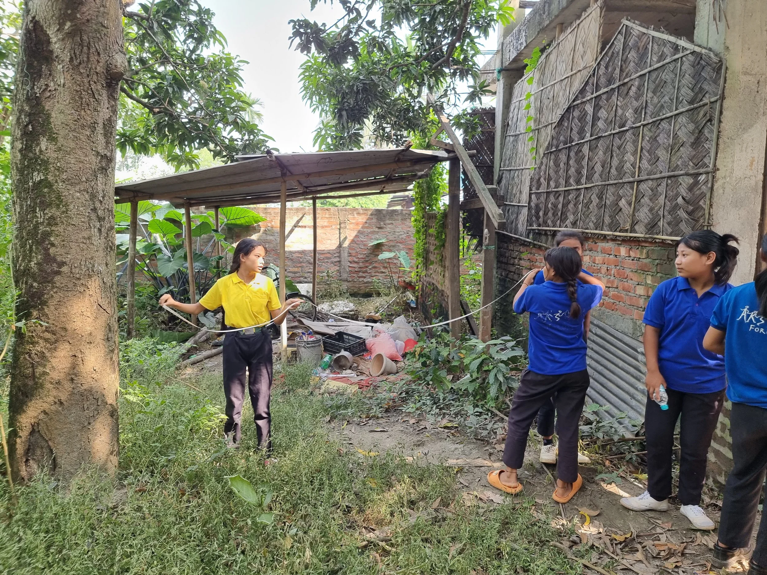 Group of young women in blue and yellow shirts standing and talking outdoors in a garden or backyard area with trees and a makeshift shelter.