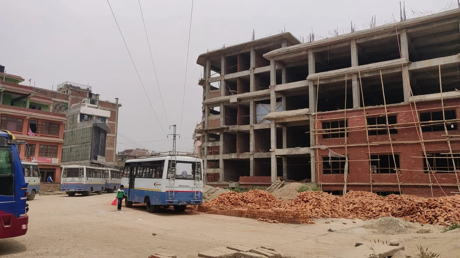 Construction site with a partially built multi-story building, construction materials, and several buses parked nearby. A person is walking on the sandy ground.