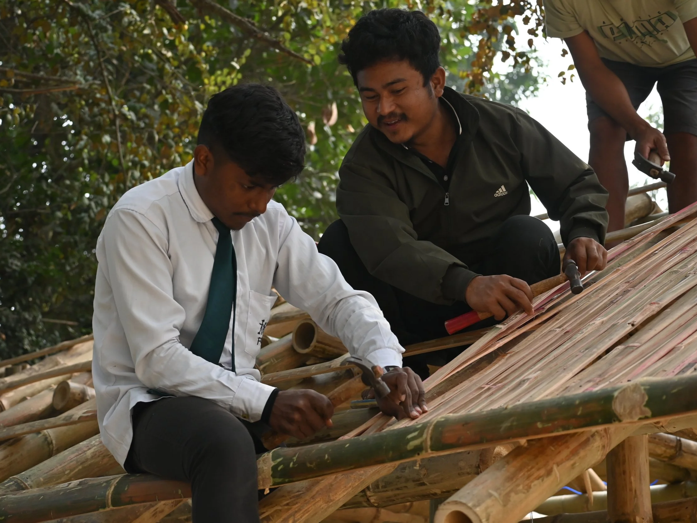 Two people working on a bamboo structure outdoors, using hammers and tools, surrounded by greenery.