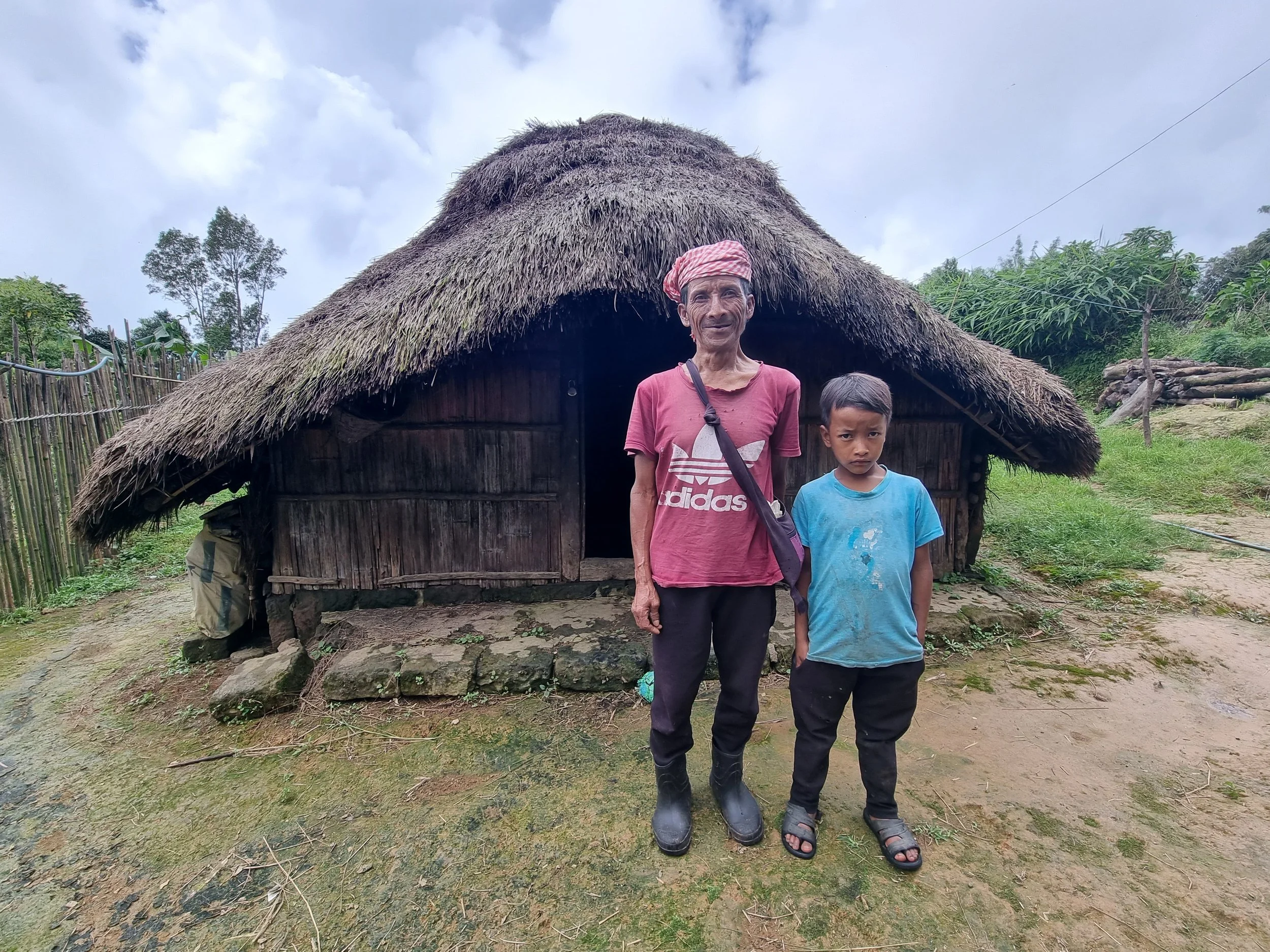 An elderly man and a boy standing in front of a traditional thatched-roof hut in a rural landscape with greenery and cloudy sky.