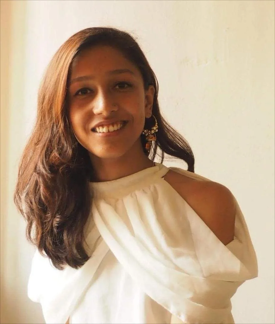 A young woman with long, wavy brown hair smiling, wearing a cream-colored off-shoulder top and gold earrings, standing against a plain background.