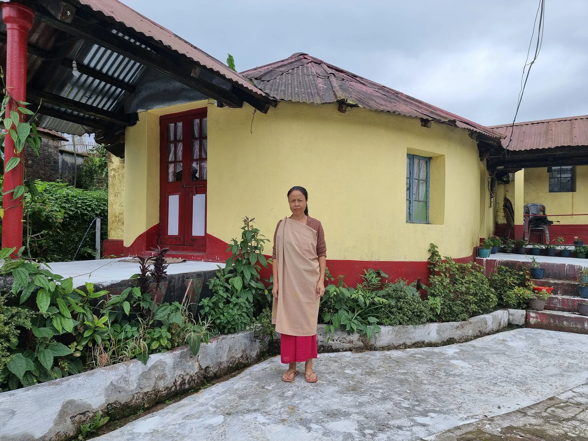 A woman standing on a concrete porch in front of a yellow house with red accents, surrounded by green plants and potted flowers. The house has a corrugated metal roof and a small porch with chairs.