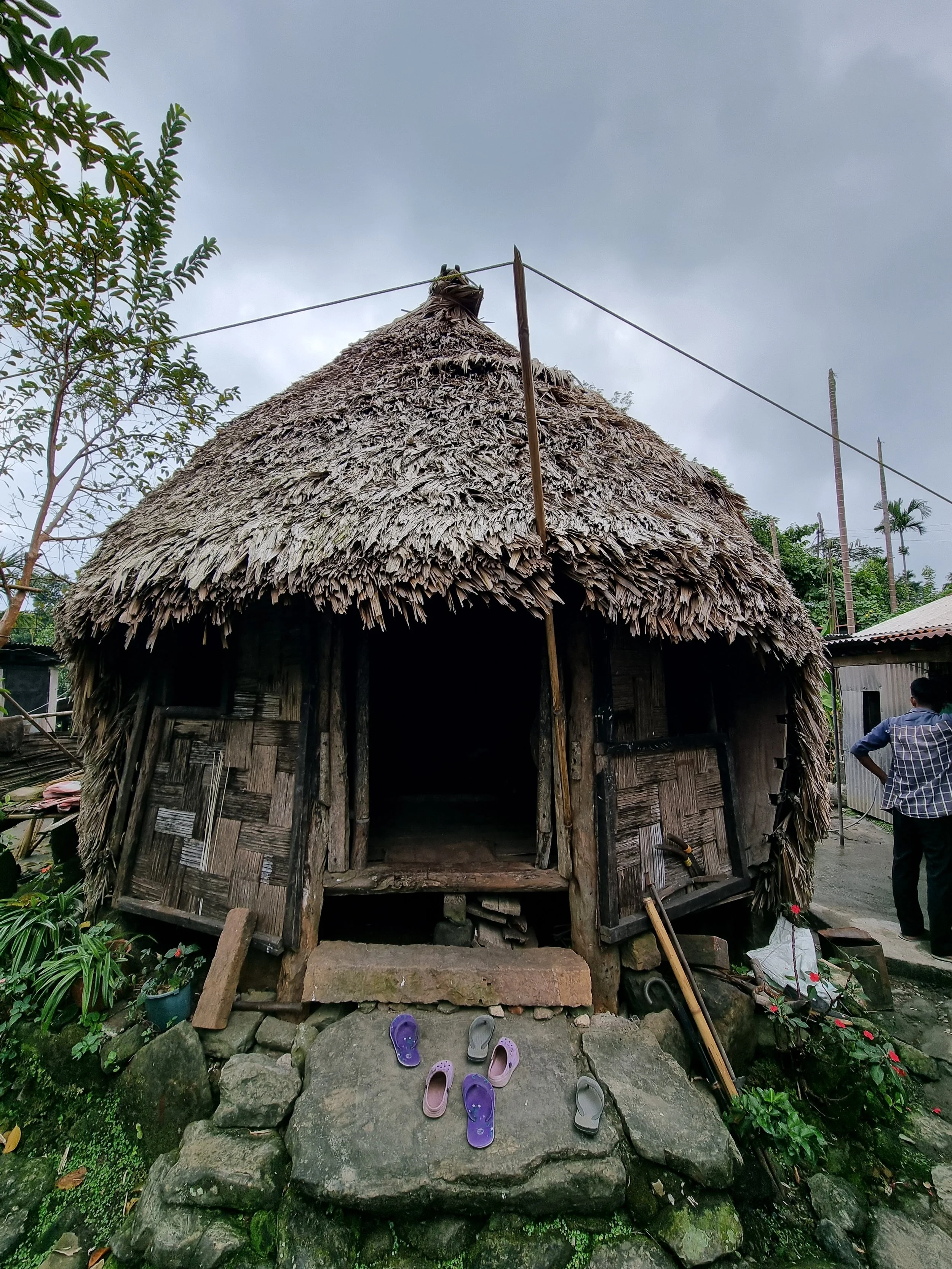 Traditional hut with a thatched roof and wooden walls, with several pairs of shoes placed on the stones at the entrance. A man stands nearby looking at the hut.