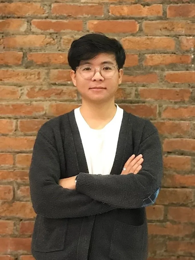 A young man with black hair and glasses standing with arms crossed in front of a brick wall, wearing a white shirt and black cardigan.