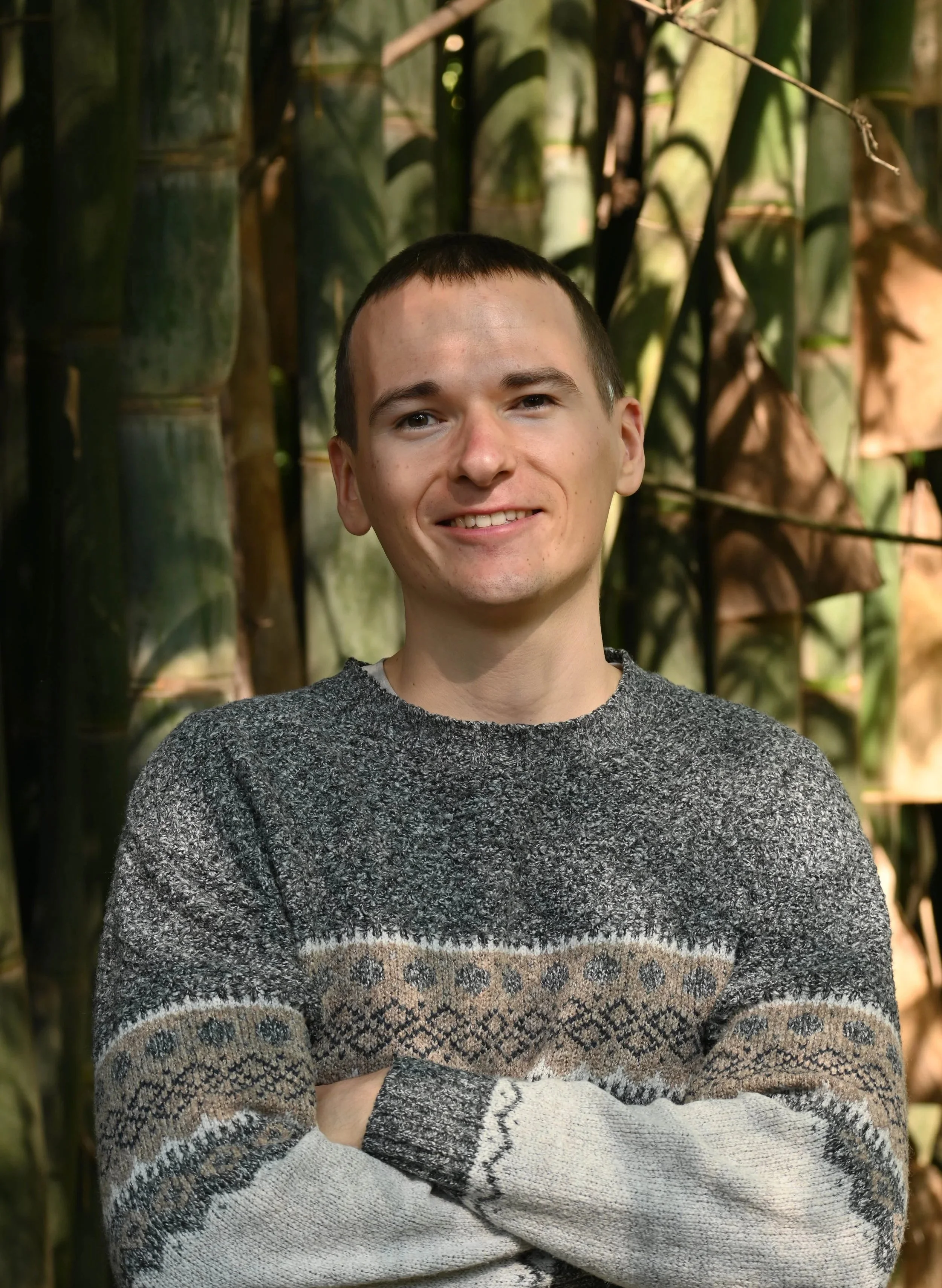 Smiling young man with short hair wearing a patterned sweater standing outdoors in front of green bamboo plants.