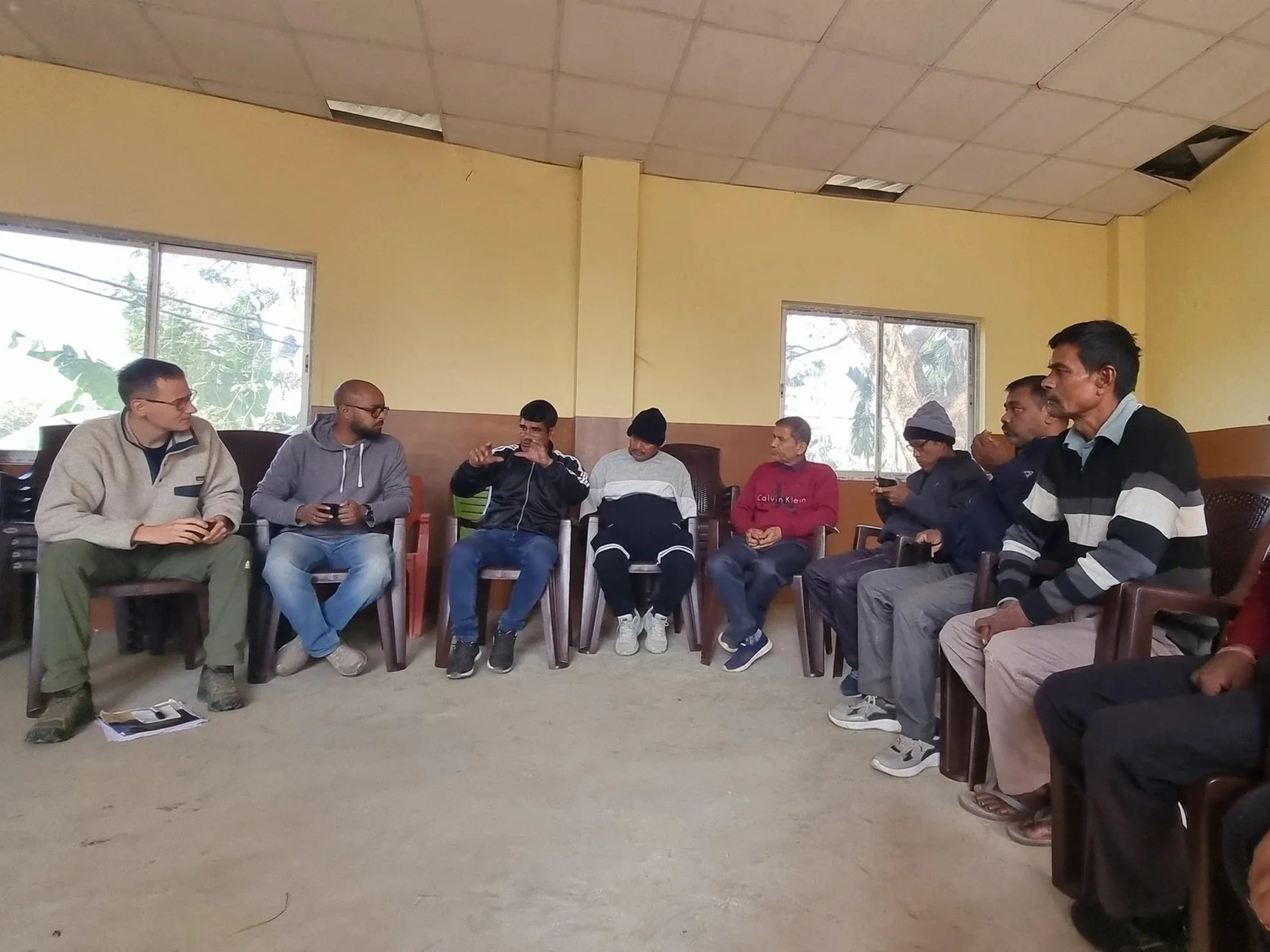 A group of eight people sitting in a room, engaged in conversation, with some holding smartphones and papers, and two windows providing natural light.