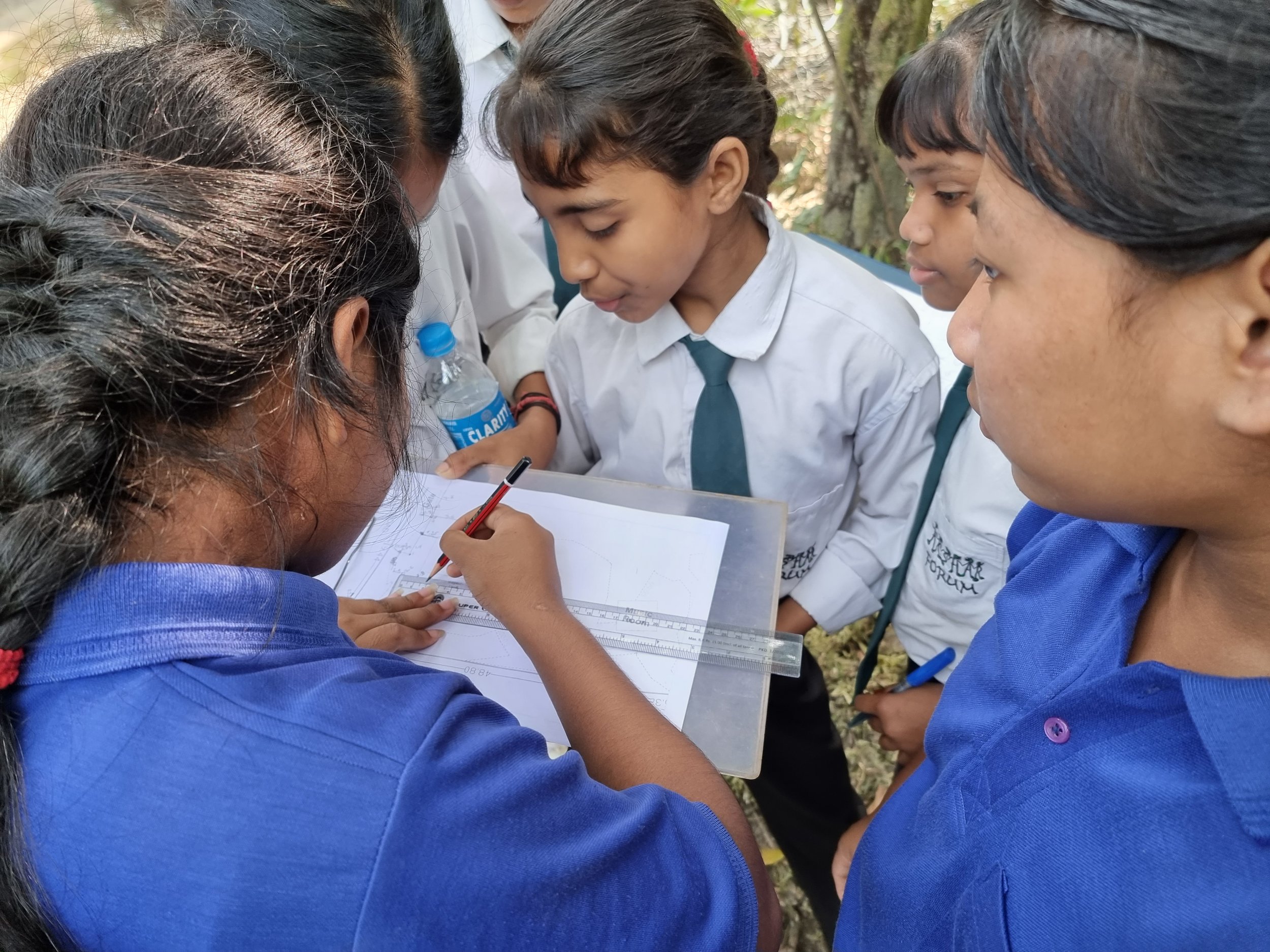 Group of school children in uniforms gathered around a woman who is explaining something on an engineering drawing or blueprint, outdoors.