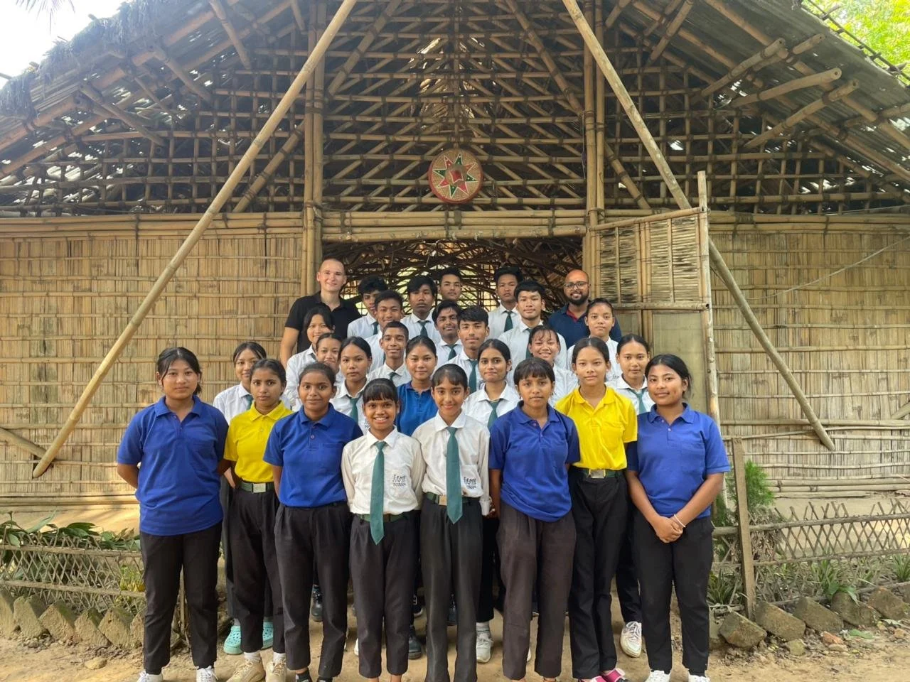 A group of school students and two adults posing for a photo in front of a bamboo structure.