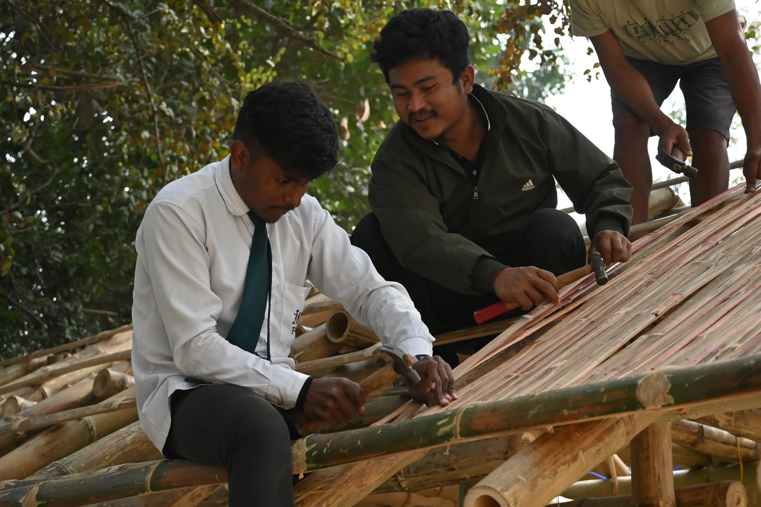 Two men working on a bamboo roof, using hammers to secure the bamboo, with another person assisting on the right, against a backdrop of trees.