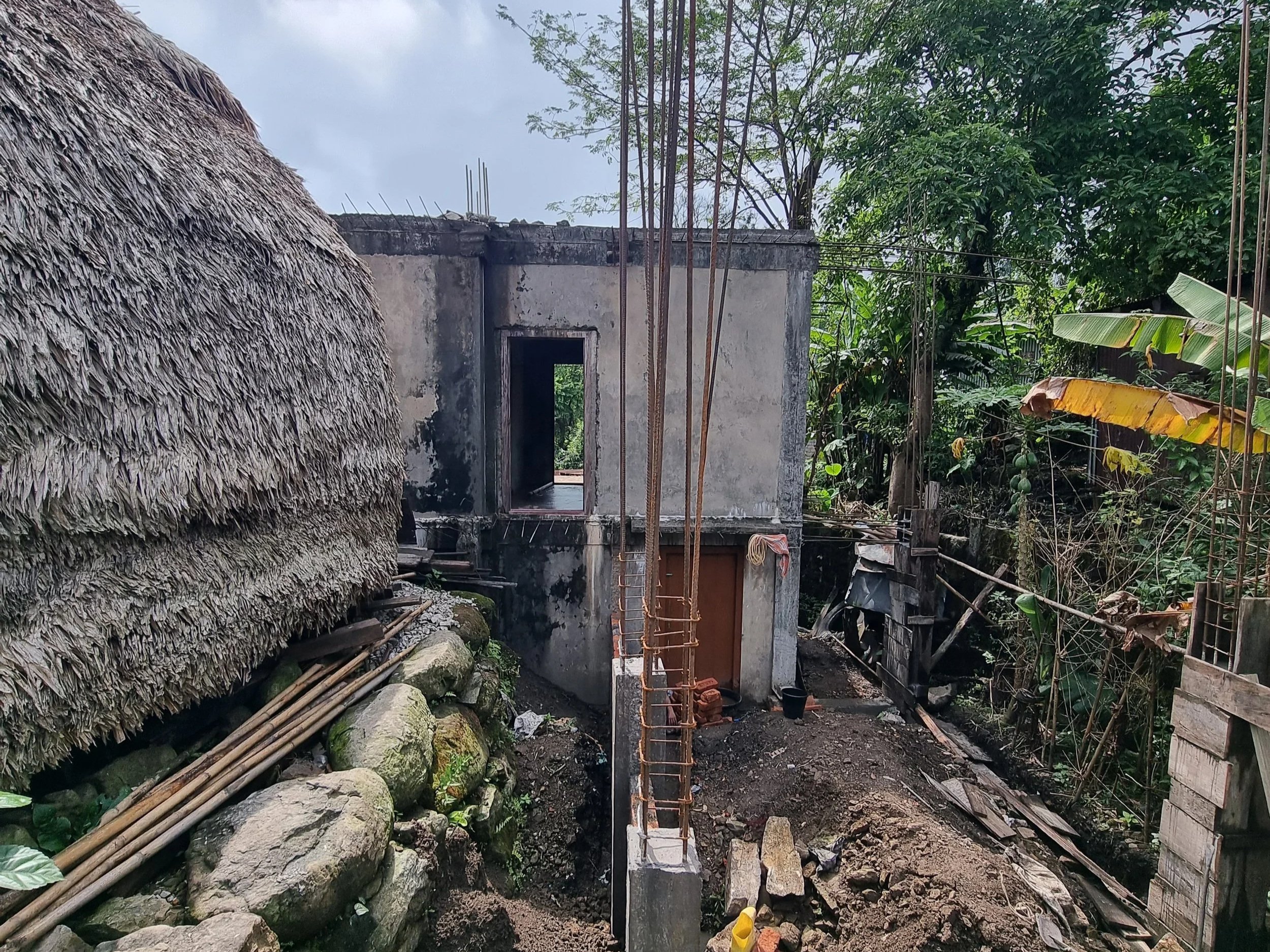 Construction site with a partially built concrete house, bamboo and metal rebar, and lush green trees surrounding it.