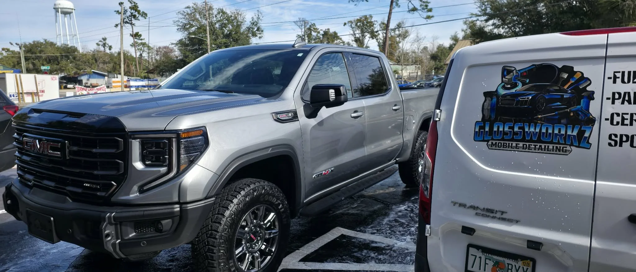 Silver GMC pickup truck parked next to a white mobile detailing van that has a logo for Glossworkz Mobile Detailing with images of car detailing tools and a sports car.