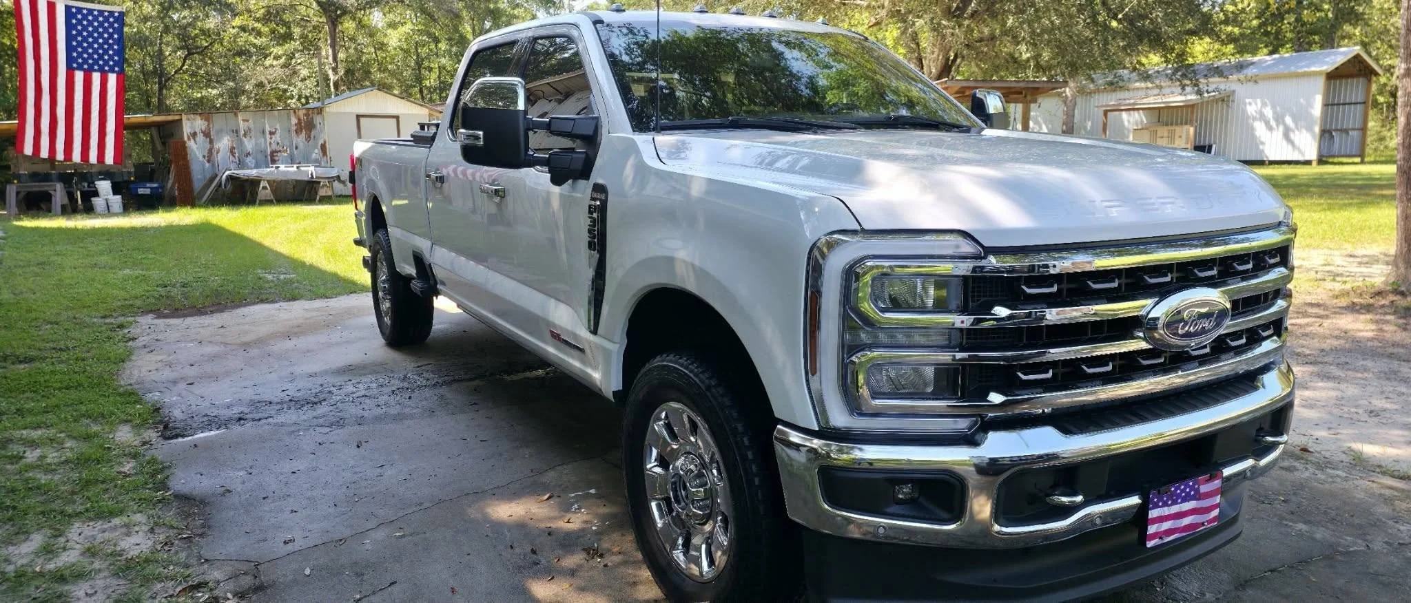 White Ford pickup truck parked outdoors on a concrete pad, with a green grassy area and trees in the background, and an American flag hanging from a pole on the left side.