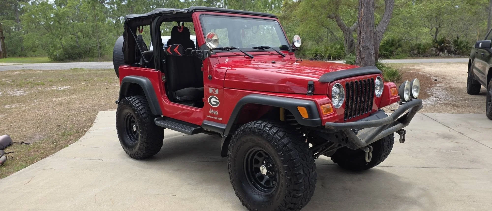 Red Jeep Wrangler with off-road tires and black accessories parked on a concrete driveway surrounded by trees.