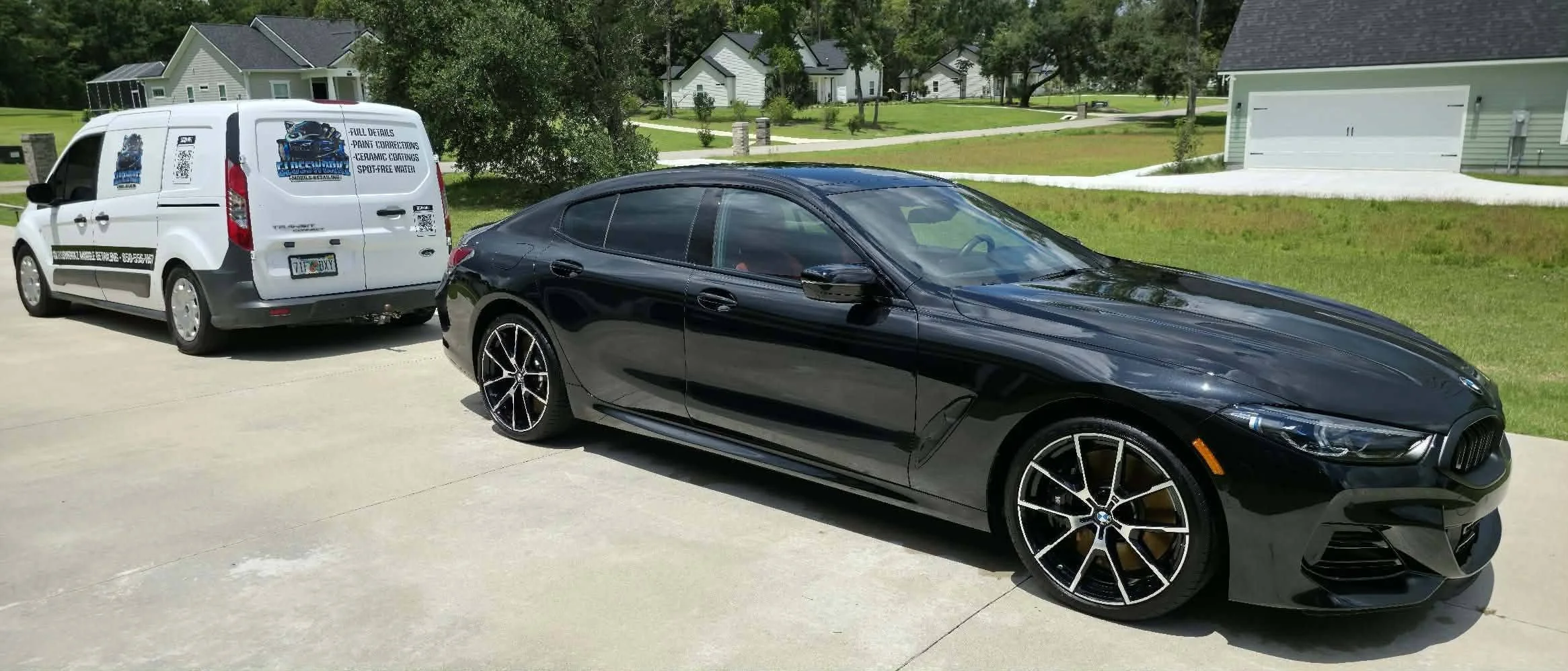 A black BMW coupe with tinted windows parked on a driveway next to a white service van with logos and text advertising ceramic coating and paint correction services, in a residential Crawfordville,FL neighborhood with houses, lawns, and trees.