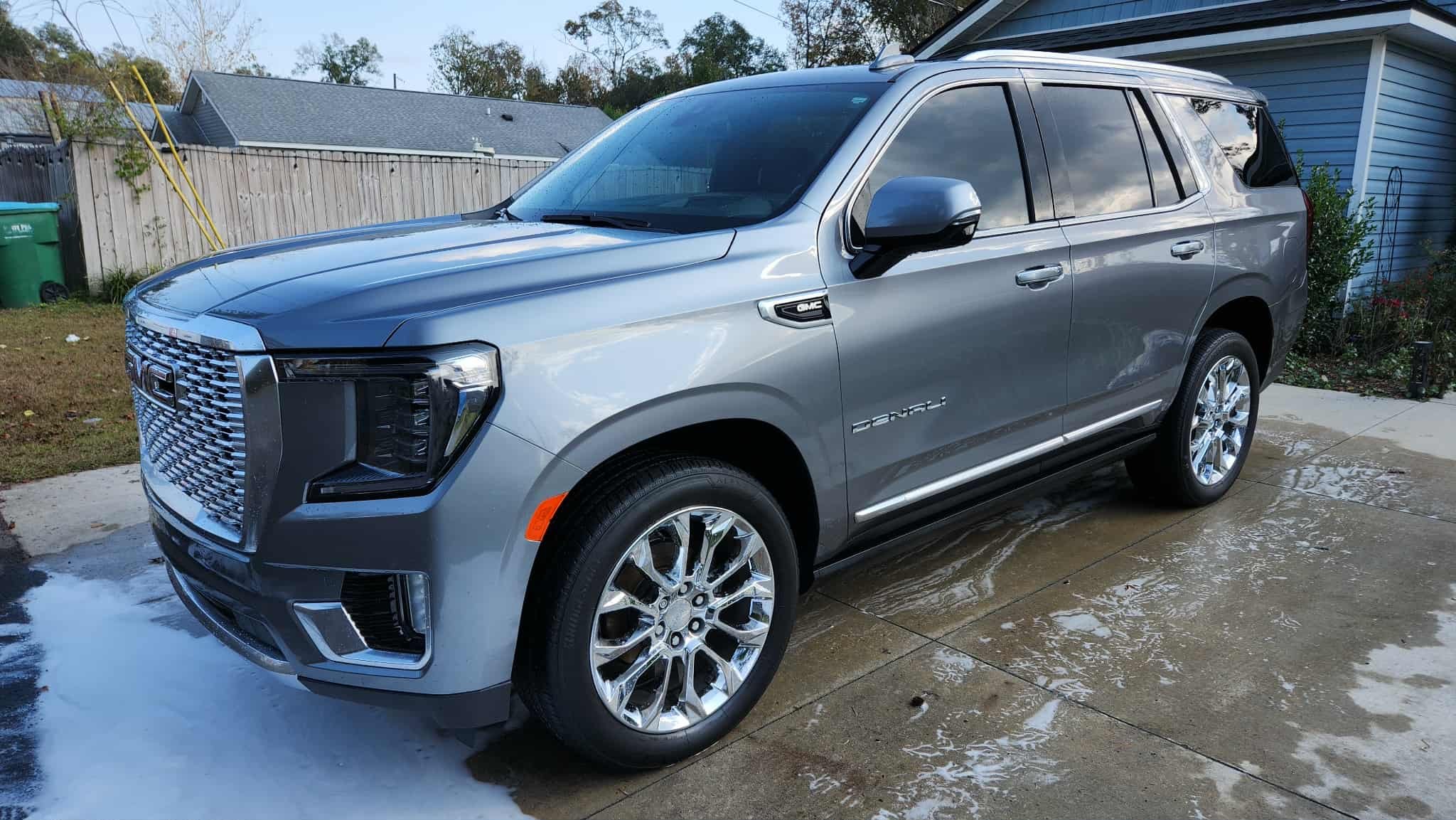 A silver GMC Yukon Denali SUV parked on a driveway after washing, with soap and water on the ground, in a residential backyard with a wooden fence, house, and trees in the background.