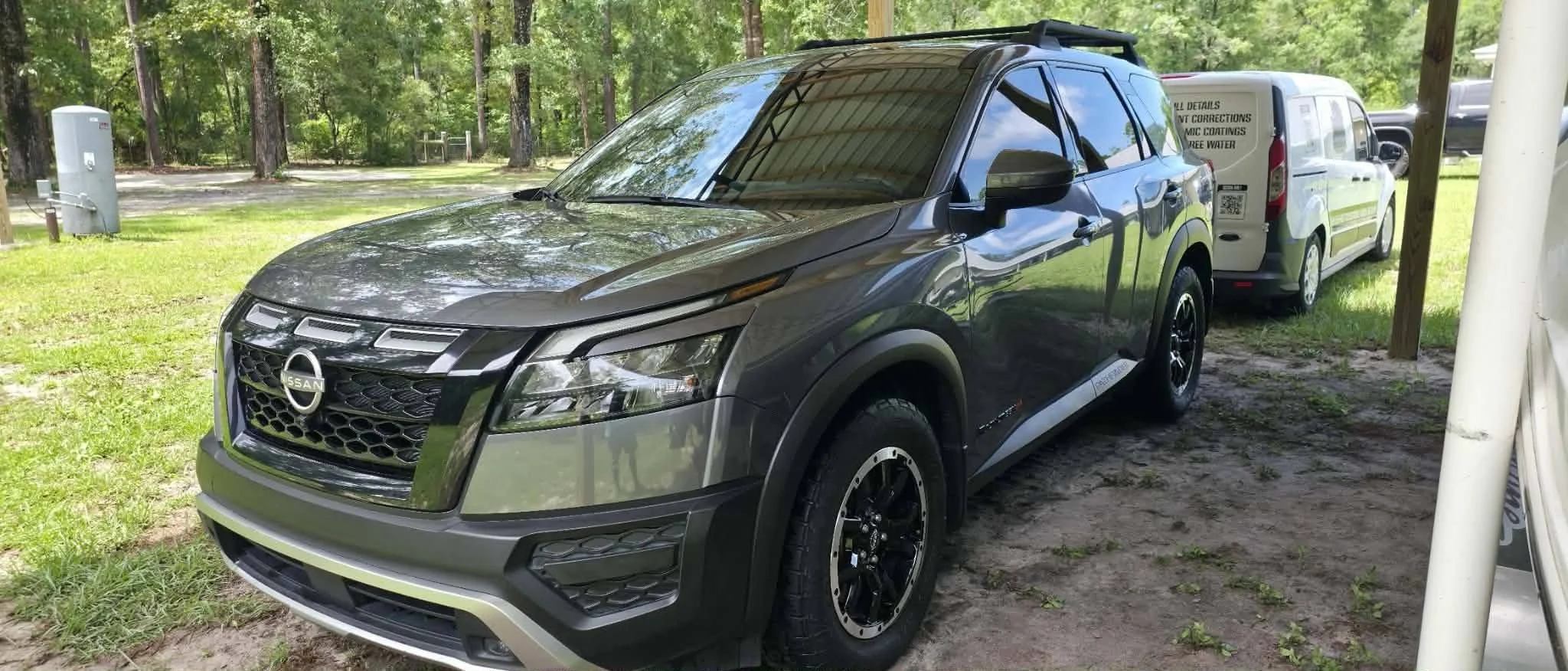 A gray Nissan SUV parked outdoors near a wooded area with a white service vehicle attached behind it.