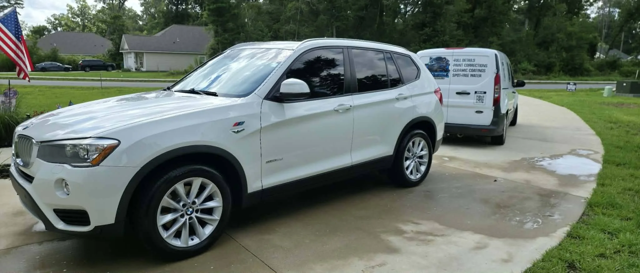 A white BMW X1 SUV parked in a Crawfordville,FL driveway with a service van behind it, in a suburban 32327 neighborhood with houses and trees in the background.