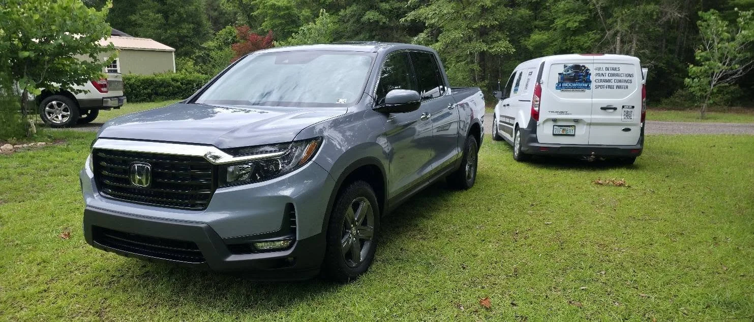 A silver Honda Ridgeline truck parked on grass with a white van and another vehicle in the background, surrounded by trees and a small house.
