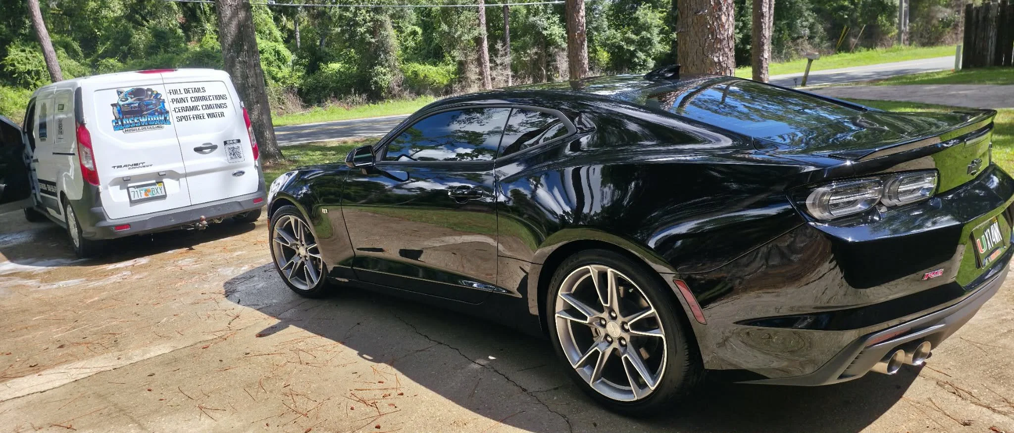 A black sports car Camero parked next to a white work van on a driveway surrounded by trees in Wakulla County, FL.