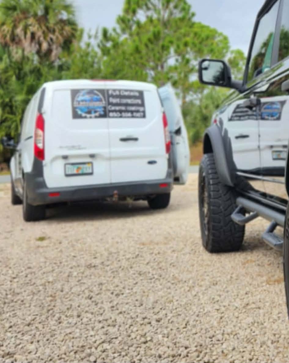 Two parked vehicles on a gravel surface, one white cargo van and one black truck, with trees in the background. The van has a business sign on the back and the truck has large tires. Truck belongs to woman and fur babies