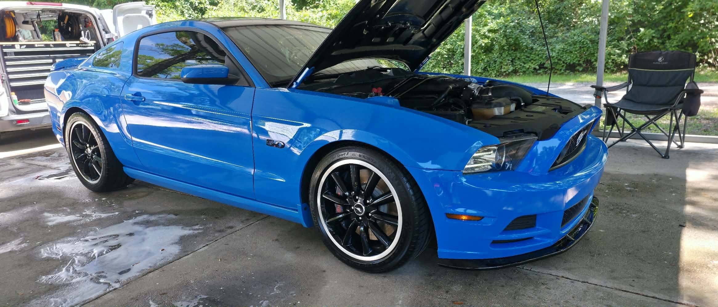 Blue Ford Mustang with opened hood in a carport, next to a black folding chair and a toolbox in the background.