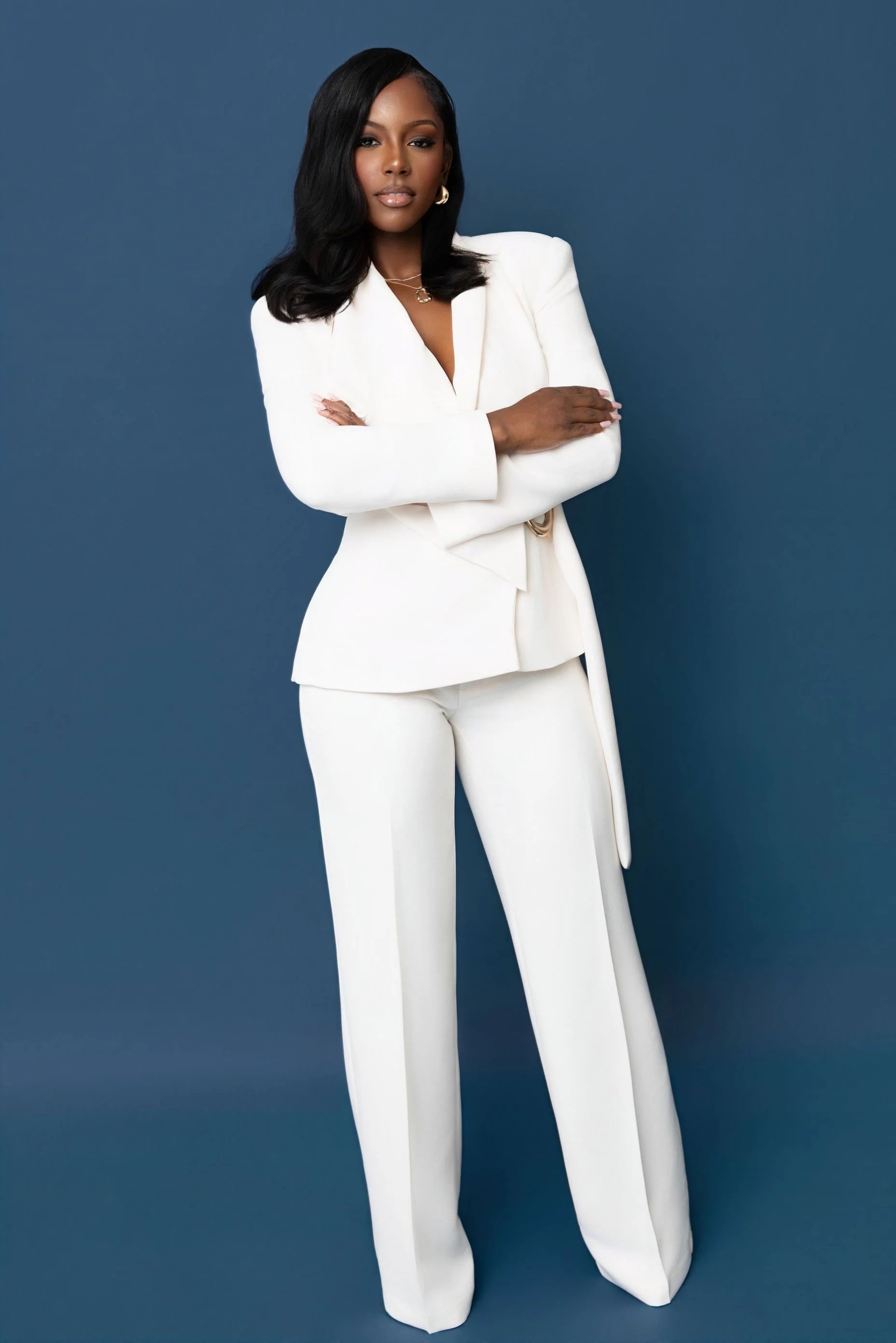 A woman in a white suit standing with arms crossed against a dark blue background.
