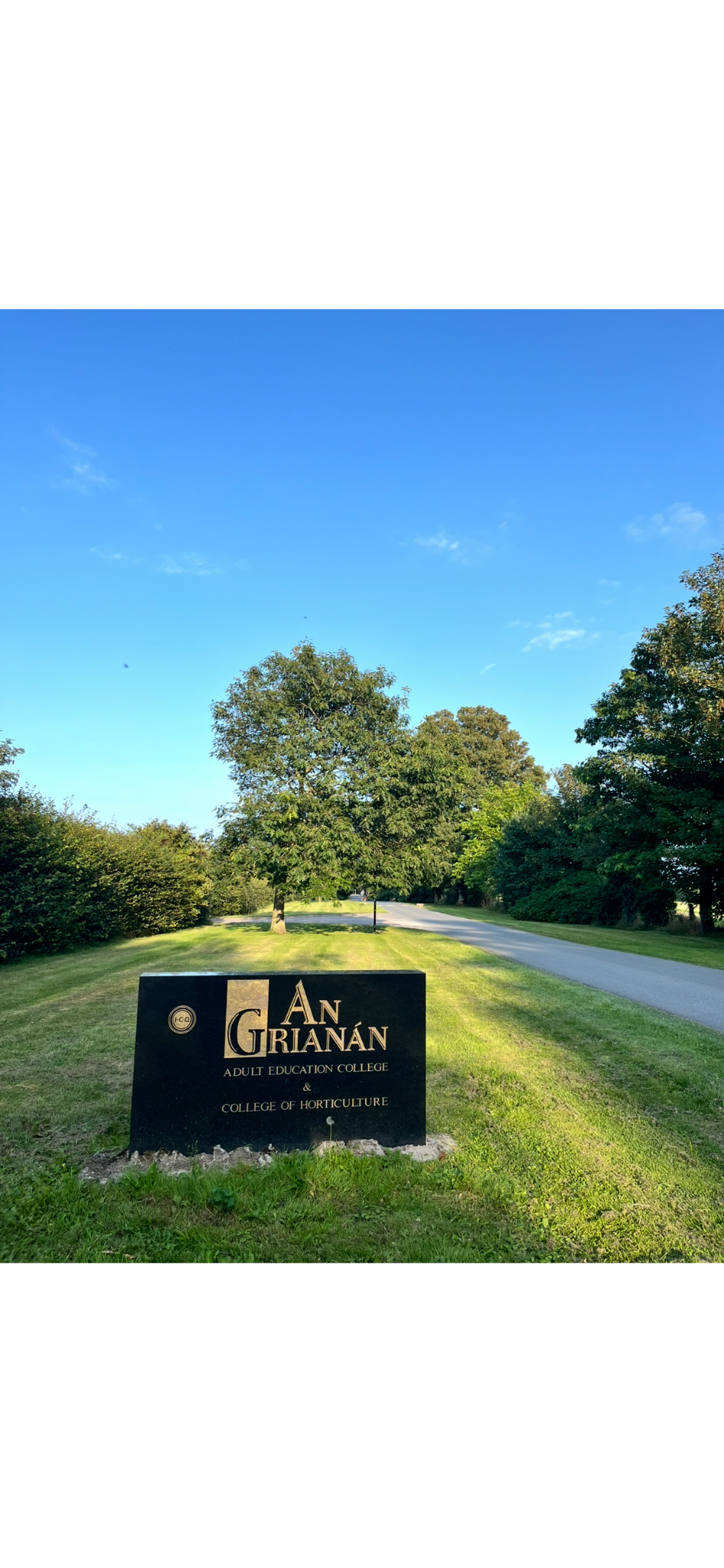 Sign for An Grián Adult Education College and College of Horticulture at a park with a grassy lawn, trees, and a clear blue sky.
