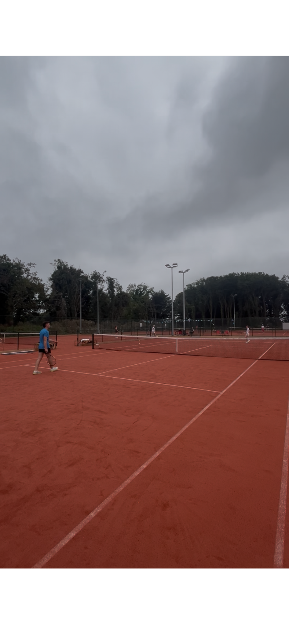 A cloudy day at an outdoor tennis court with players practicing and walking around.