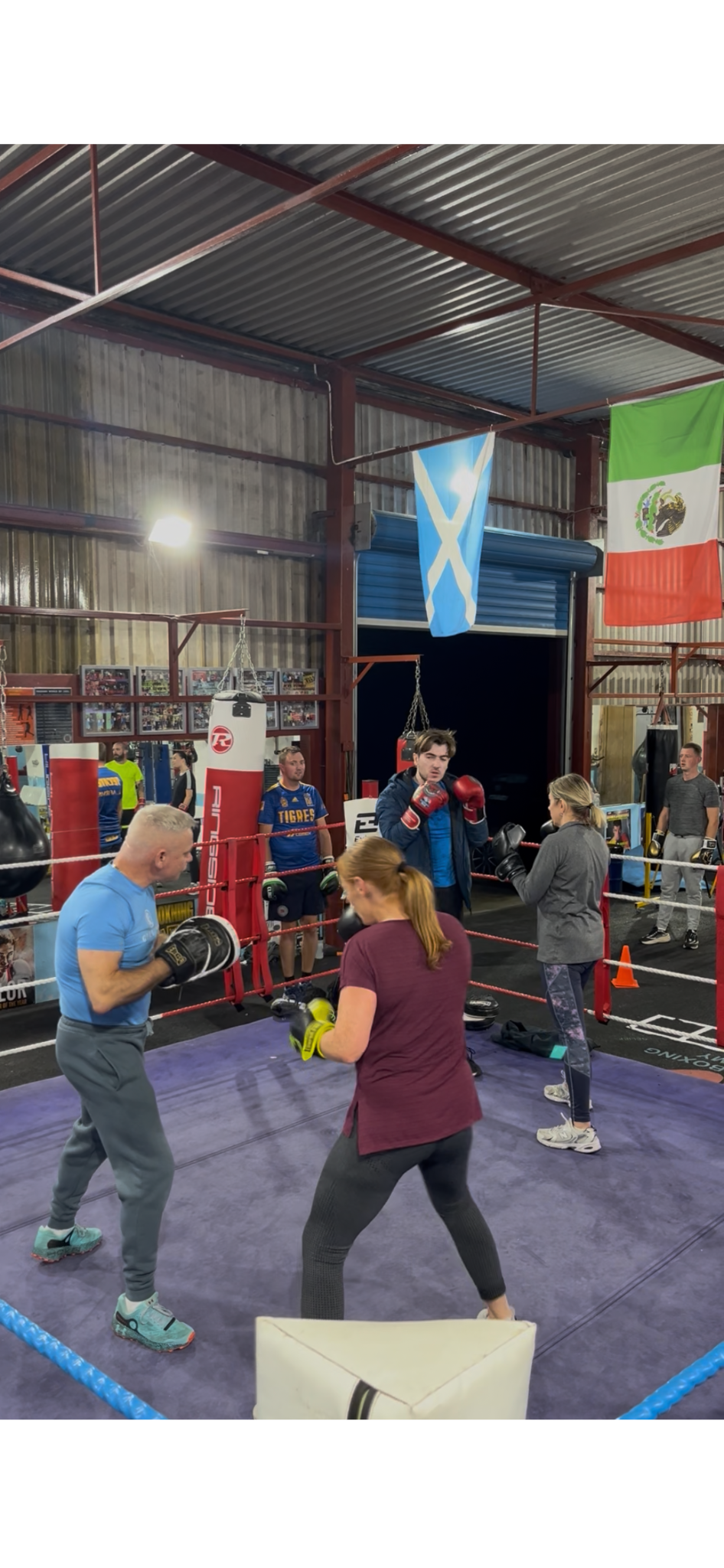 People practicing boxing in a gym with punching bags, flags hanging from the ceiling, and gym equipment around.