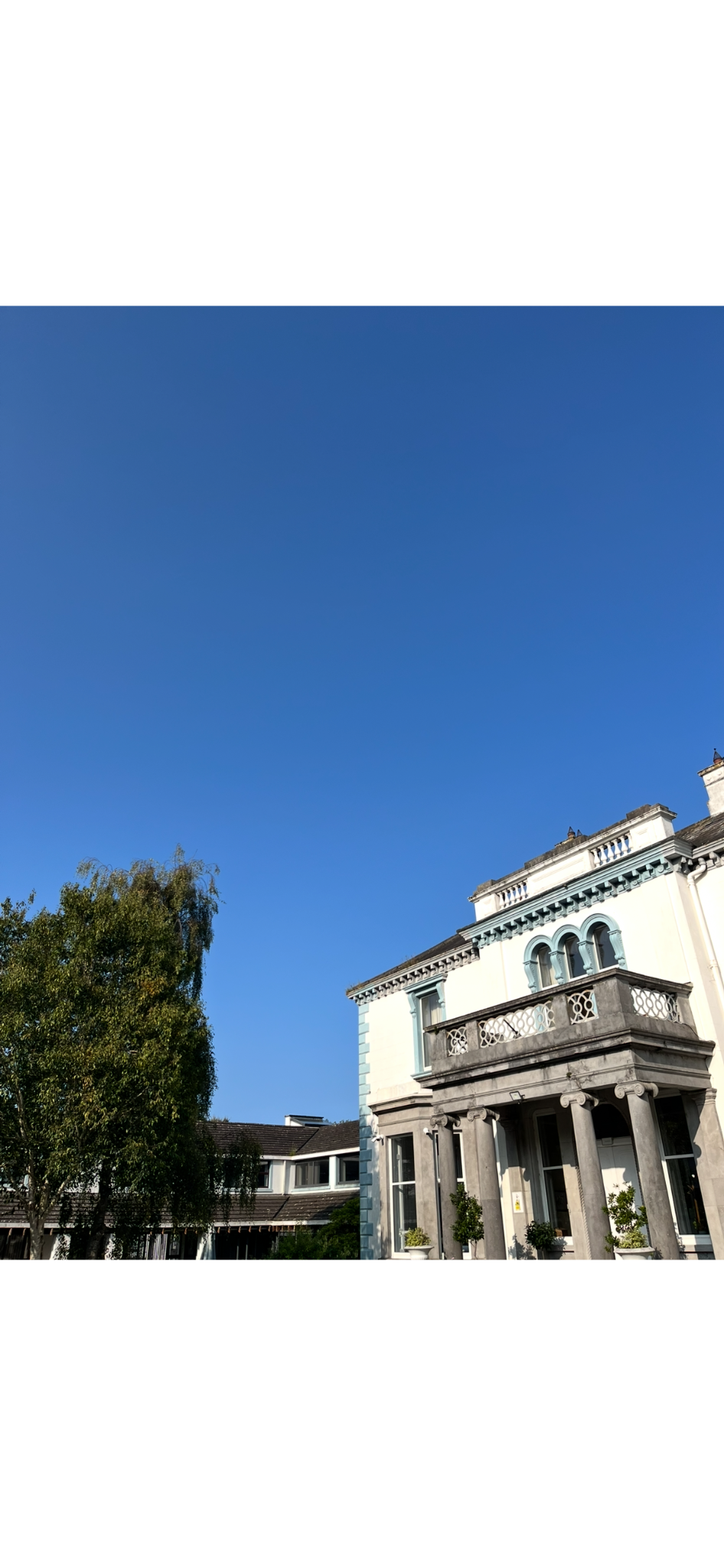 A white and gray house with classical architecture, columns at the entrance, balcony, and decorative elements, set against a clear blue sky and trees.