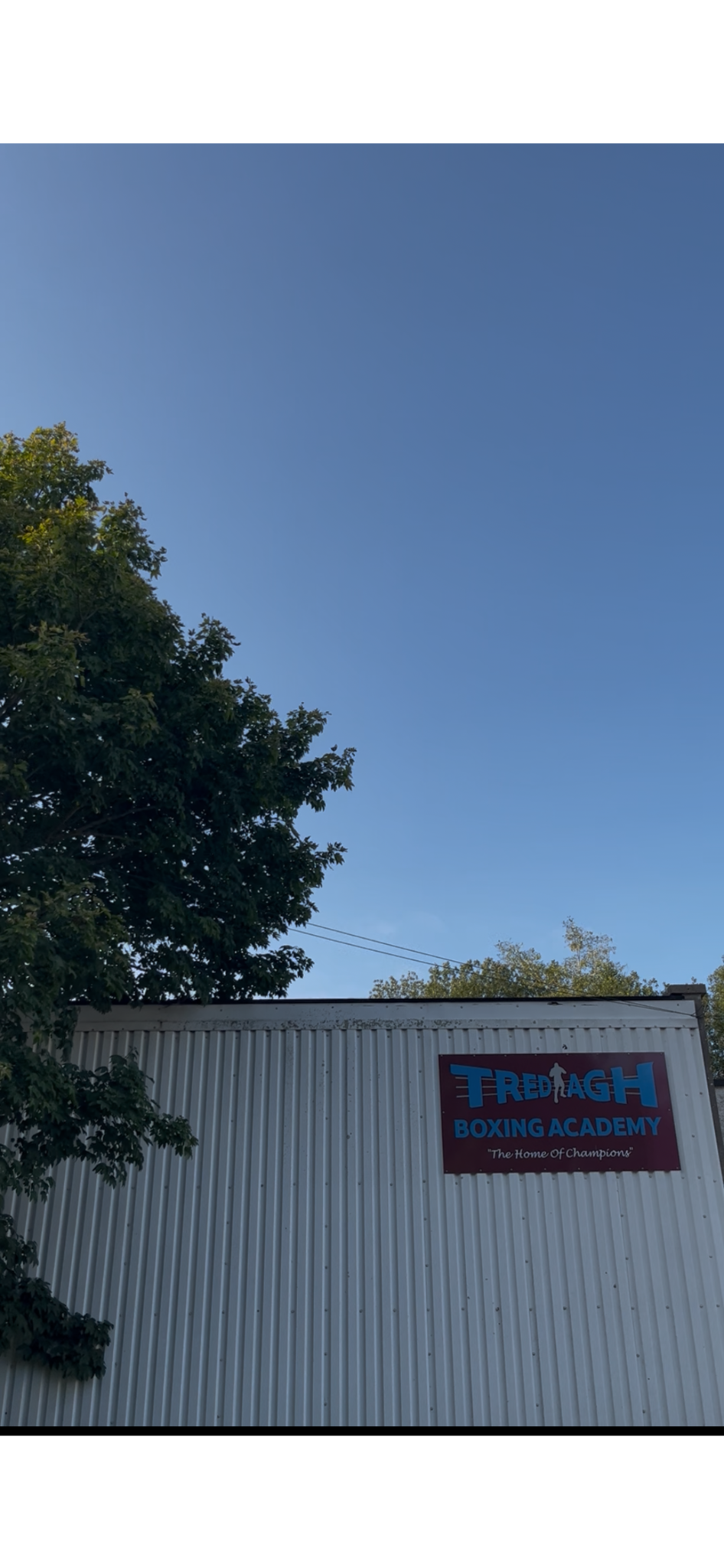 Sign for Tredagh Boxing Academy on a metal building exterior with trees and a clear blue sky in the background.