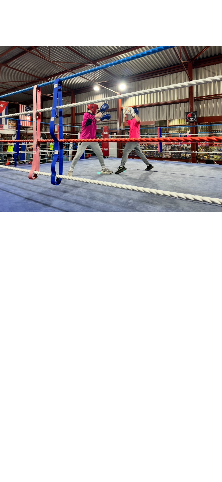 Two young boys in boxing gloves and helmets sparring in a boxing ring inside a gym.