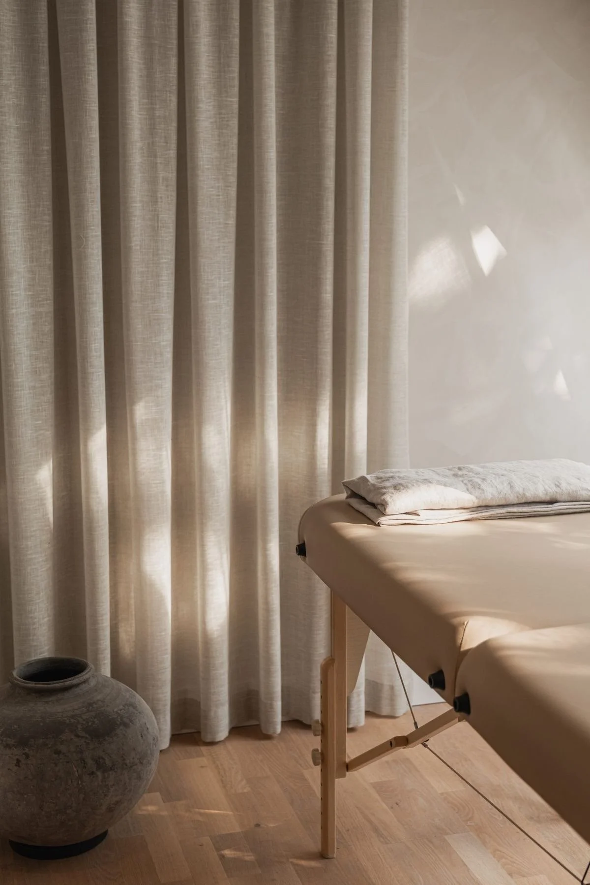 Una stanza con tende beige chiuse, un letto da massaggi con lenzuola beige e una coperta di fiandra, una grande ansa di ceramica grigia sul pavimento di legno.