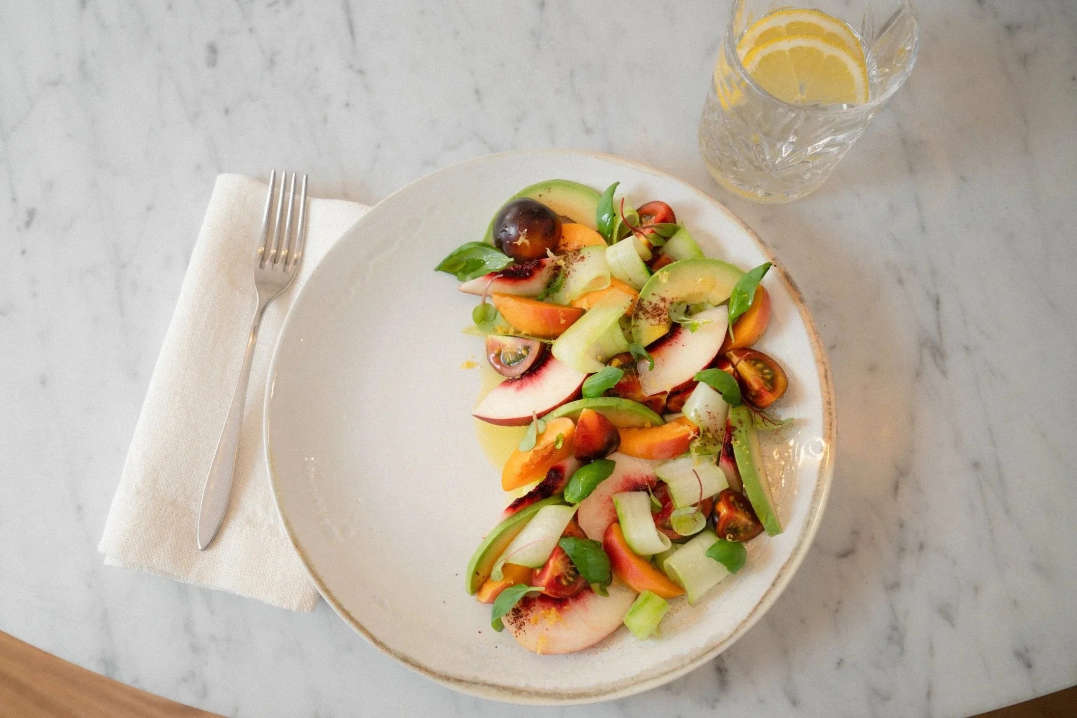 Insalata di verdure fresche e frutta, con pomodorini, cetrioli, fette di frutta e foglie di basilico, servita su un piatto bianco. Bicchiere con limoni a fette e acqua gassata, posato su un tavolo di marmo. Posate e tovagliolo accanto.